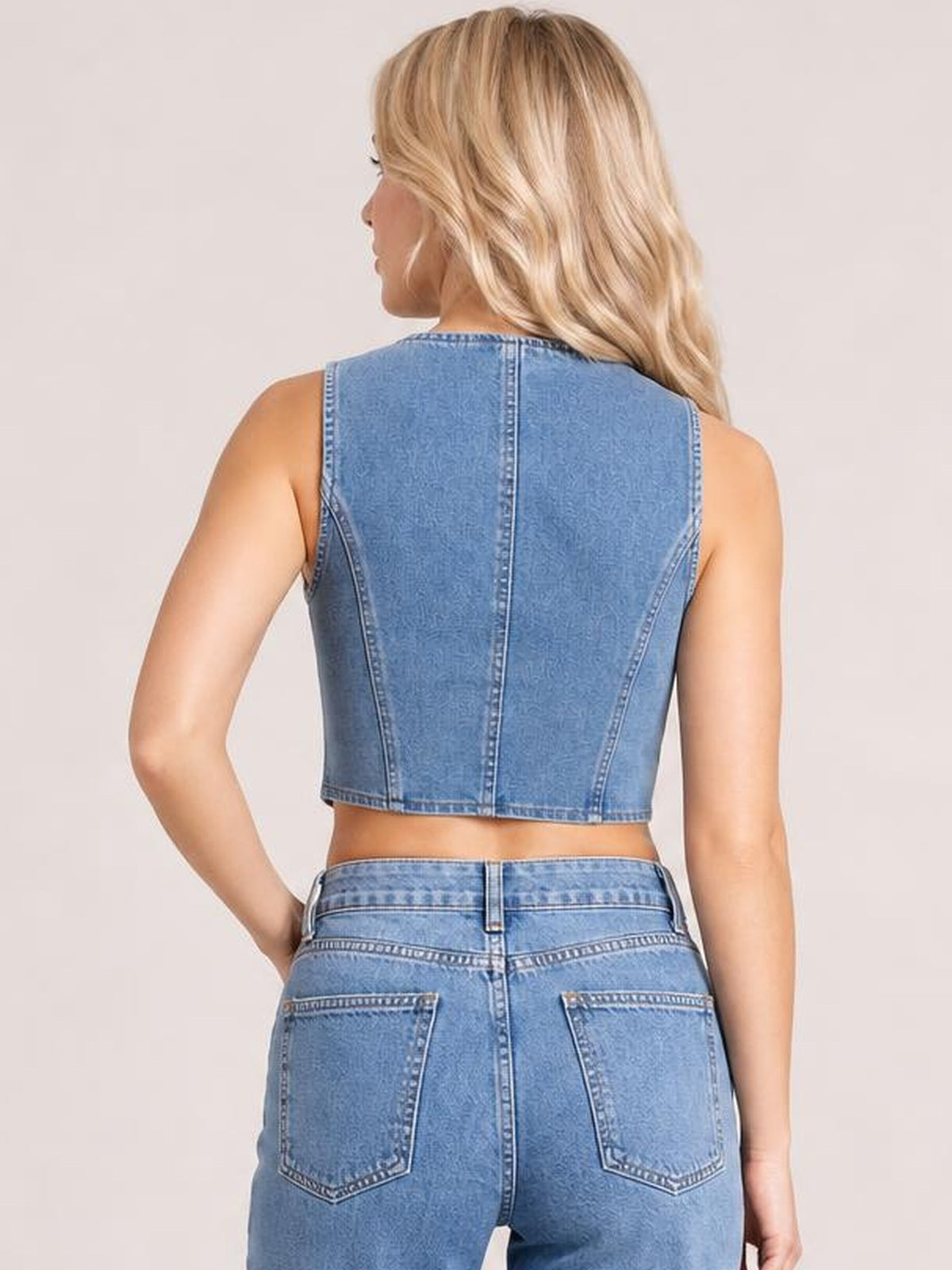 A model poses from behind in a blue denim vest with a sleeveless, cropped fitted silhouette and visible center-back and side panel seams.