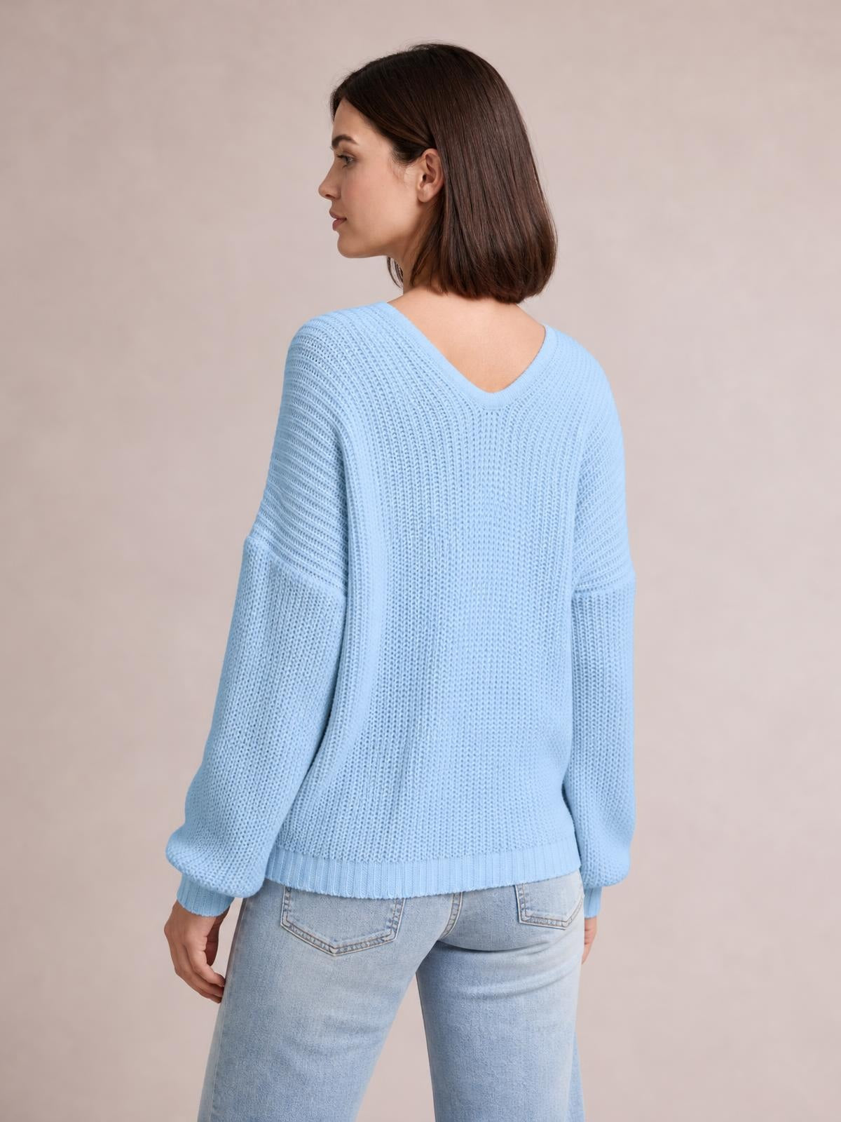 A model poses turned away in a light blue V-neck drop shoulder sweater with a relaxed silhouette, long sleeves, ribbed cuffs, and hem.
