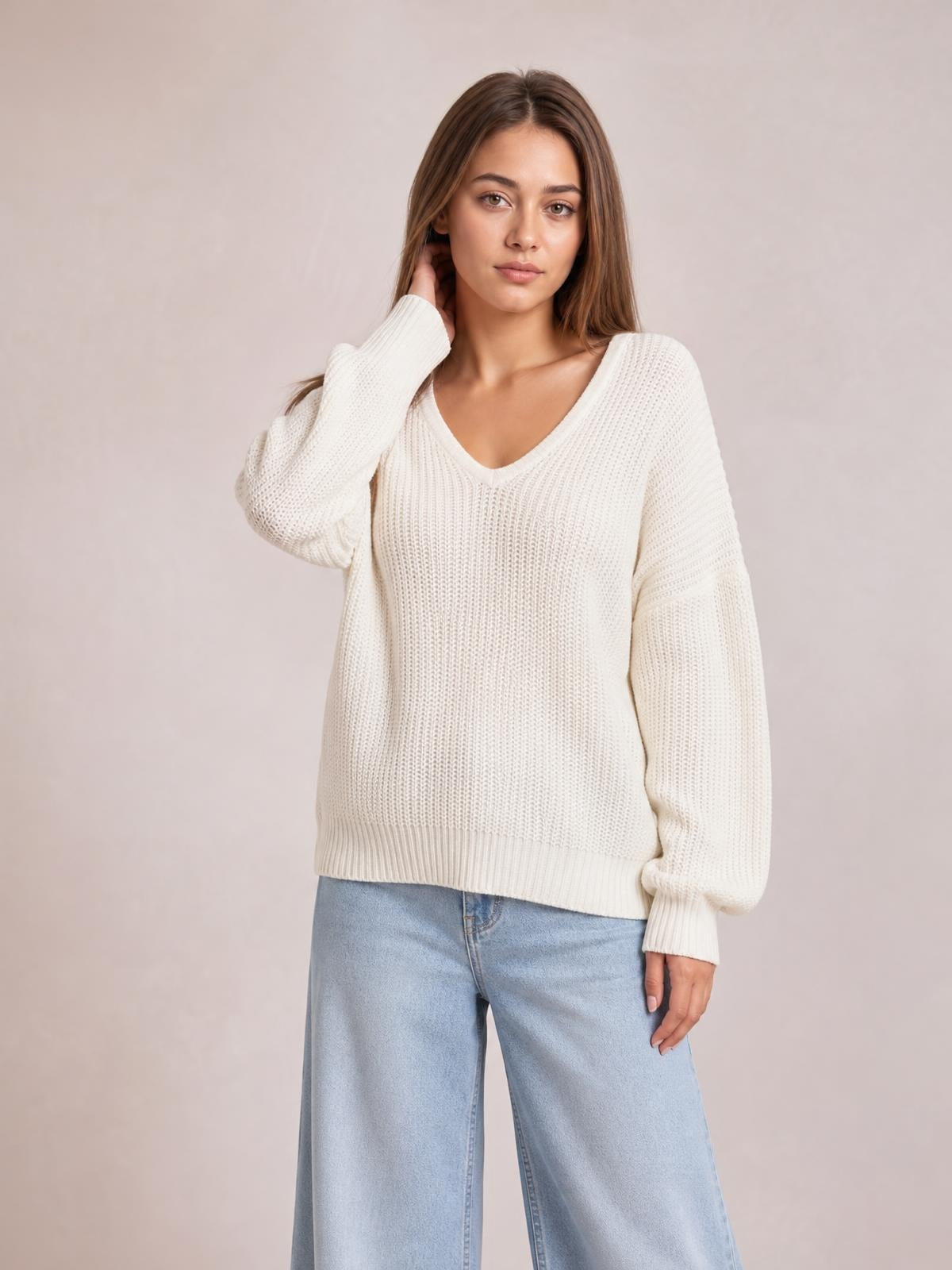 Model wearing an ivory V-neck drop shoulder sweater with a relaxed silhouette, textured knit, ribbed cuffs and hem, posing with one hand at her neck.