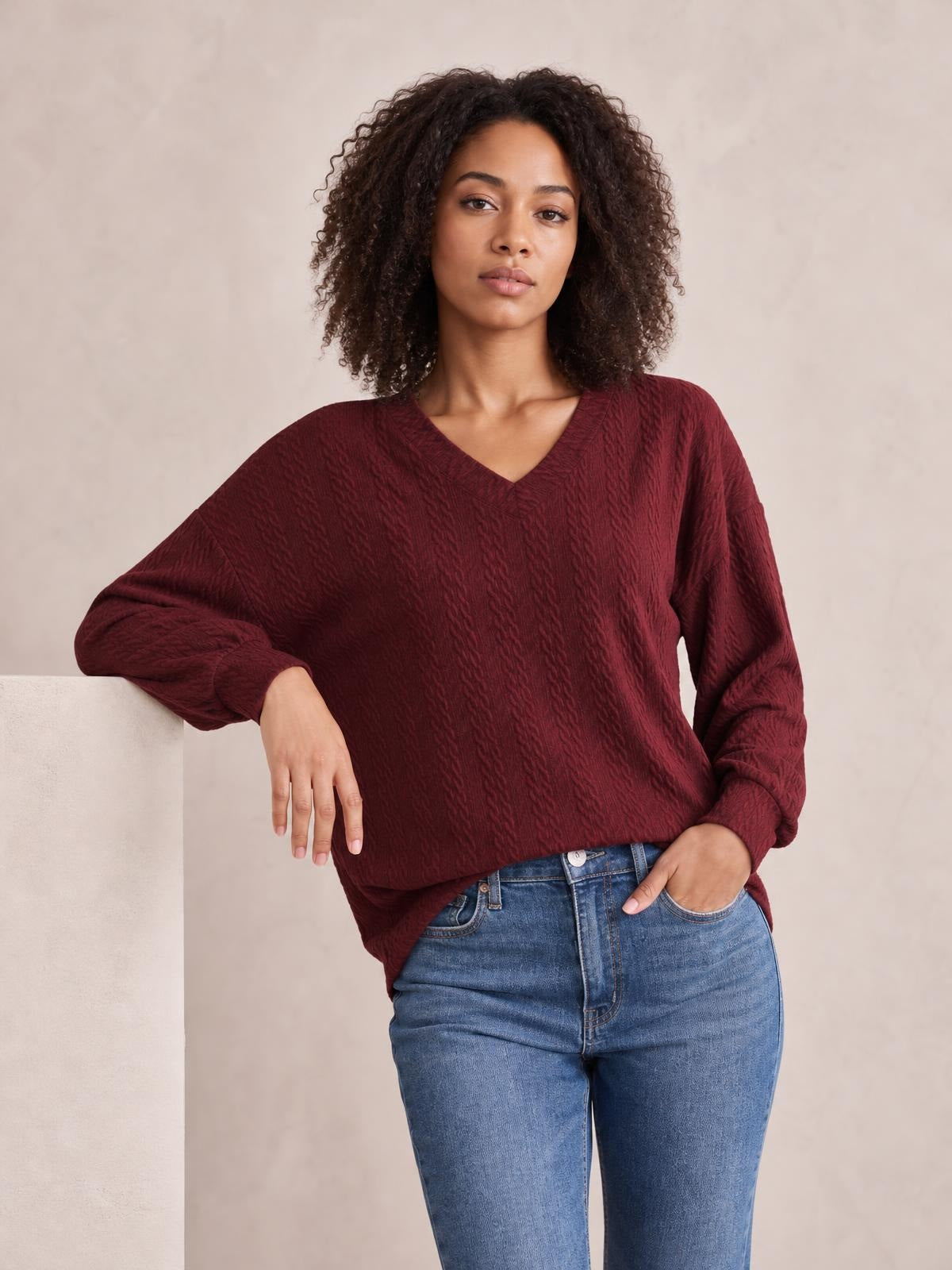Model wears a solid burgundy V-neck dropped shoulder blouse with a relaxed long silhouette, textured fabric, and long regular sleeves, posing with one hand in her jeans pocket.