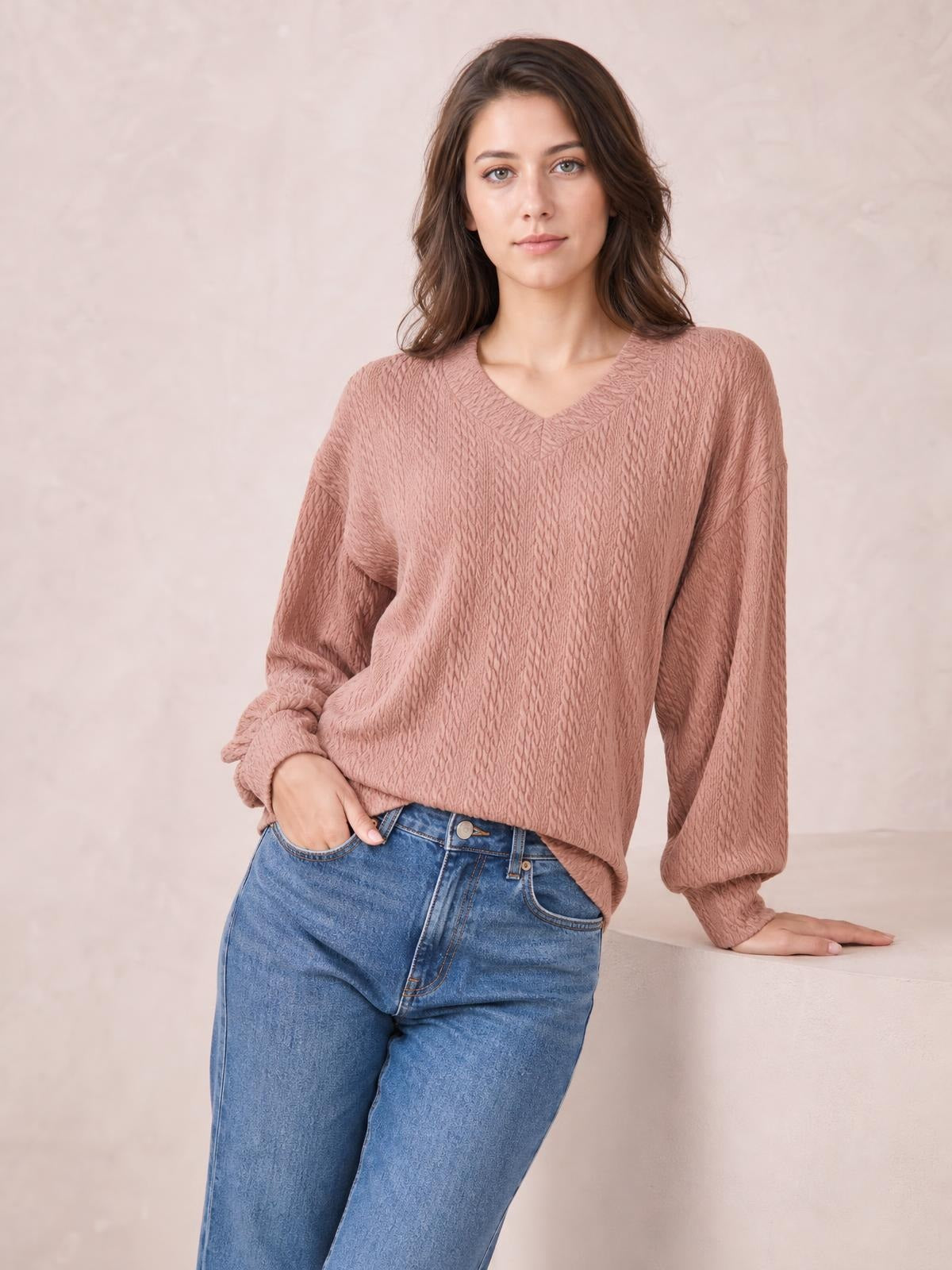 A model wears a dusty rose V-neck blouse with relaxed dropped shoulders, long sleeves, a loose long silhouette, and subtle textured construction while leaning casually.