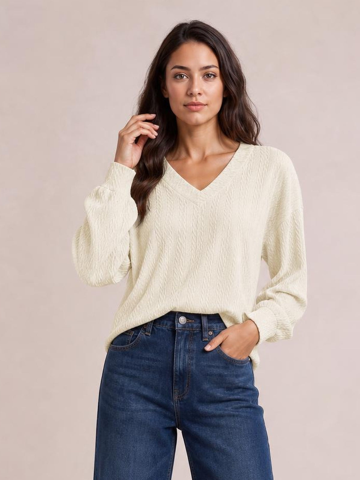 Model poses in a cream V-neck dropped shoulder blouse with a long relaxed silhouette, textured solid fabric, and long sleeves over dark jeans.