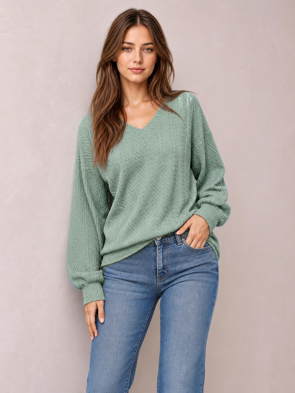 A model wears a sage green V-neck dropped shoulder blouse with long sleeves, a relaxed long silhouette, and subtle raised texture, posing with one hand in her jeans pocket.