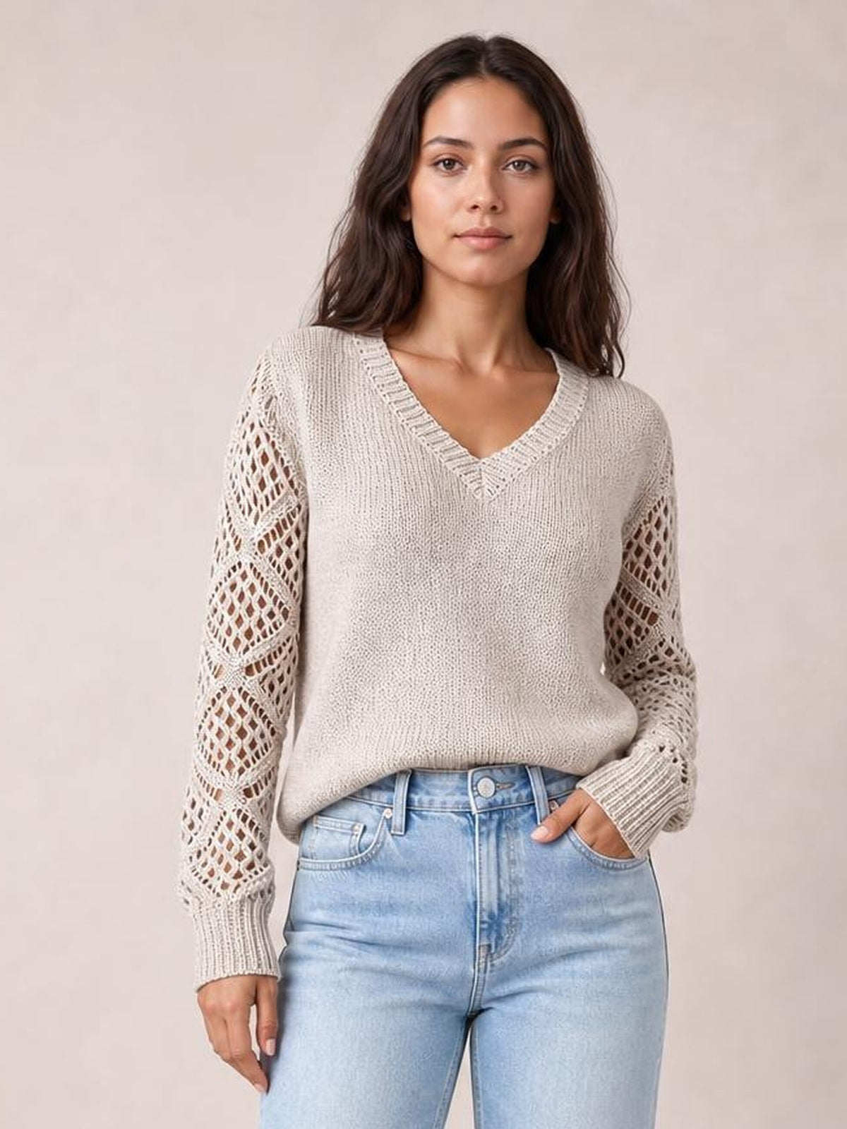 Model wears a light beige V-neck knit sweater with openwork hollow-out sleeves, ribbed cuffs, and a relaxed silhouette while standing with one hand in her jeans pocket.
