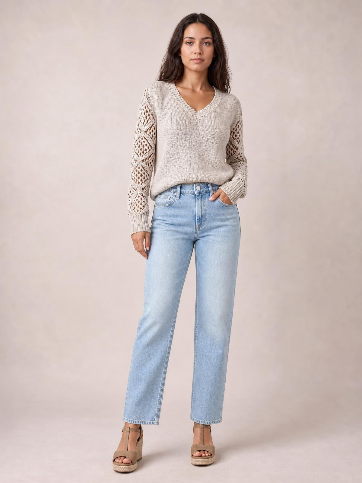Model wears a cream V-neck knit sweater with openwork hollow-out sleeves, ribbed cuffs, and a relaxed silhouette while standing with one hand in her jeans pocket.