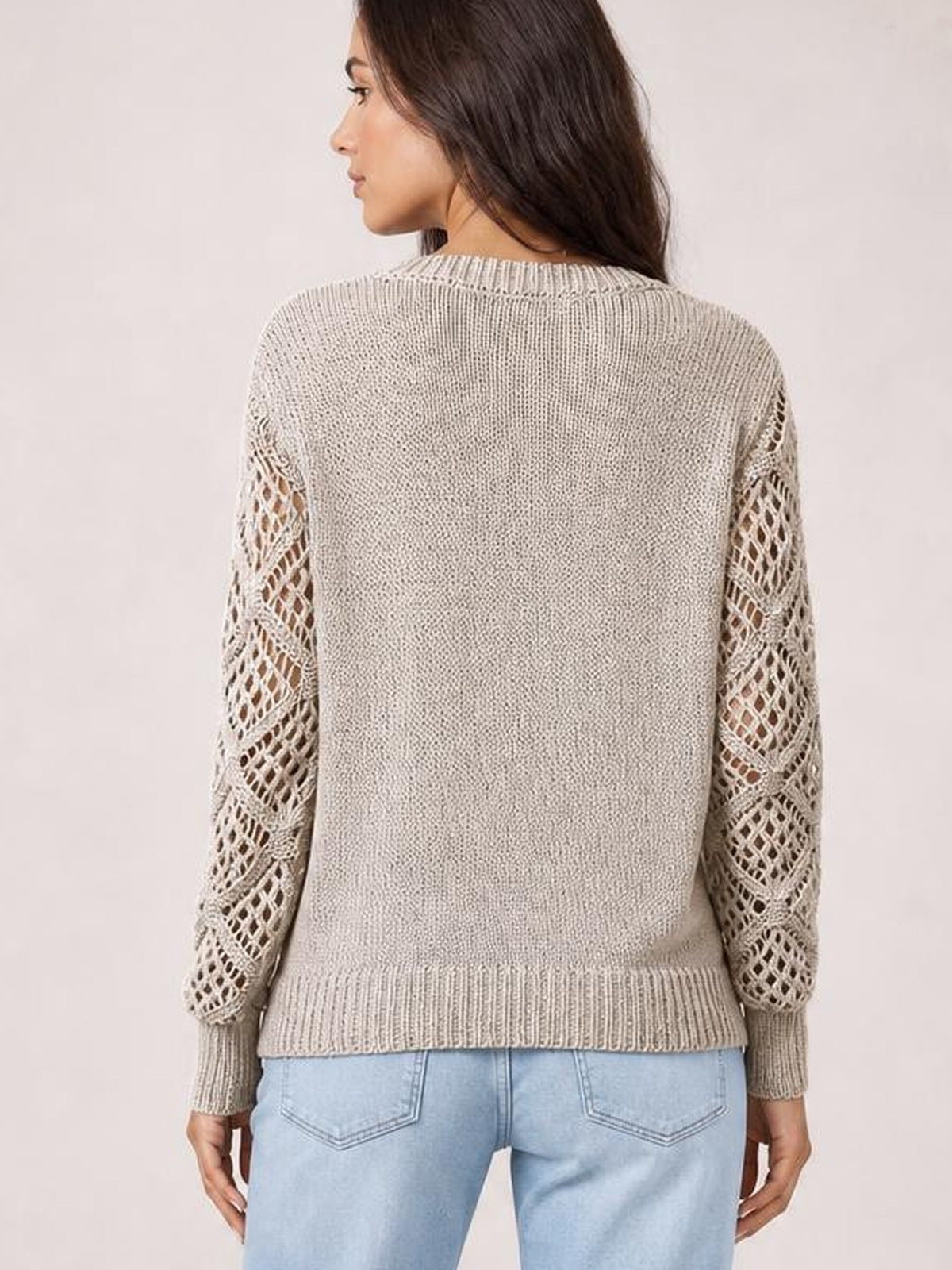 A model shown from the back wears a light beige knit sweater with openwork hollow-out sleeves, ribbed cuffs and hem, and a relaxed silhouette.