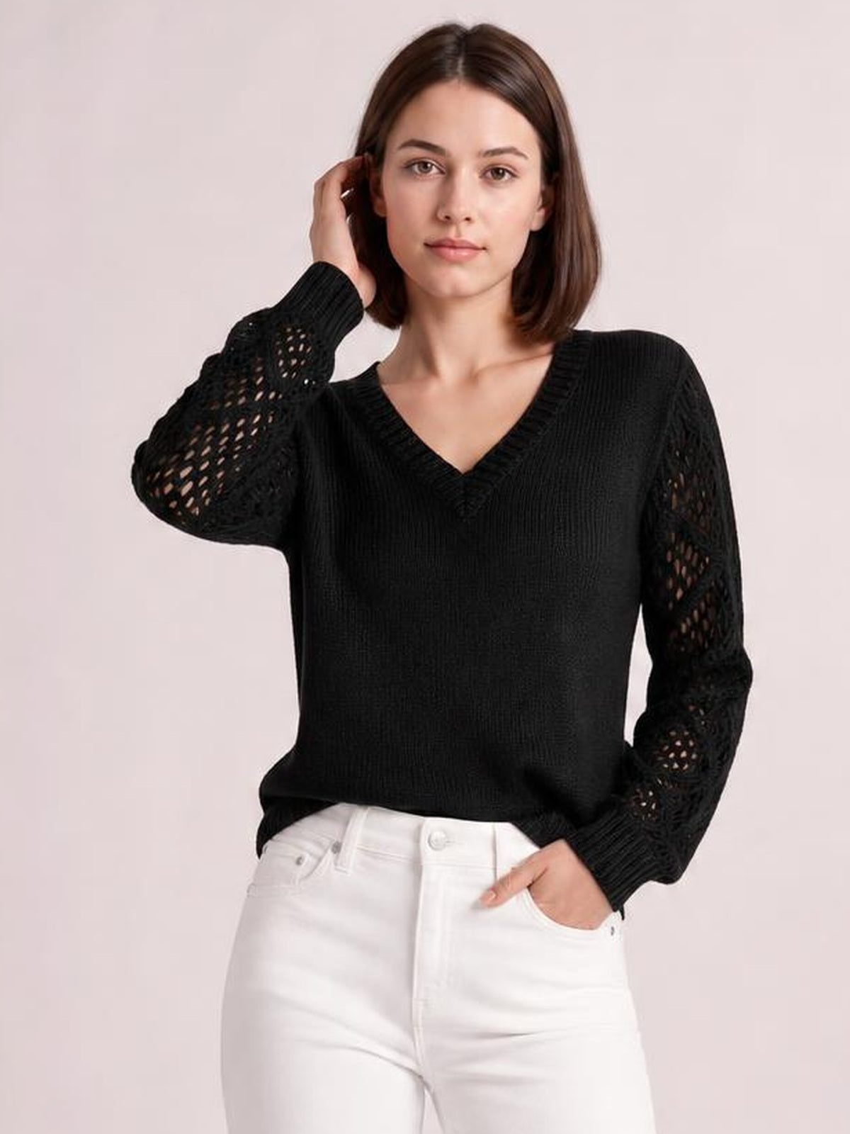 Model wearing a black V-neck knit sweater with openwork hollow-out sleeves, ribbed cuffs and hem, posed with one hand in her jeans pocket.