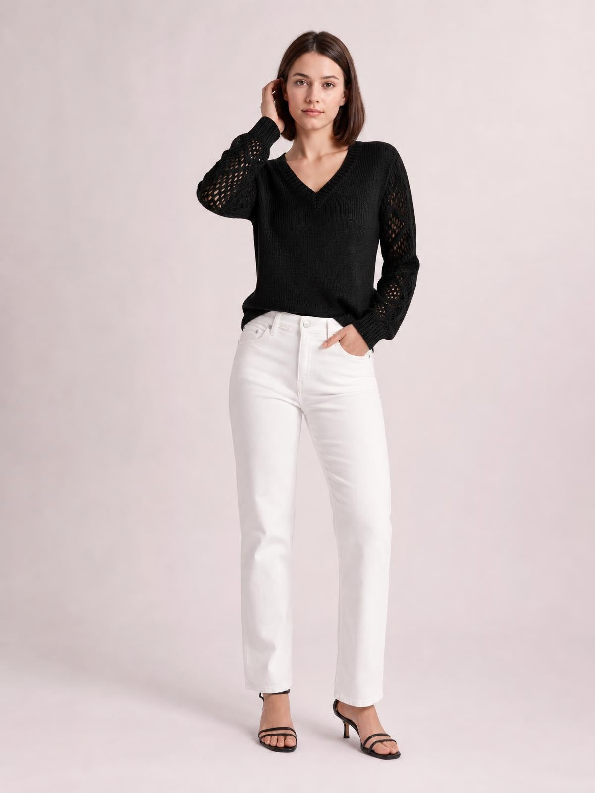 Model wears a black V-neck knit sweater with openwork hollow-out sleeves, ribbed trim, and a relaxed silhouette, posing with one hand in her pocket.