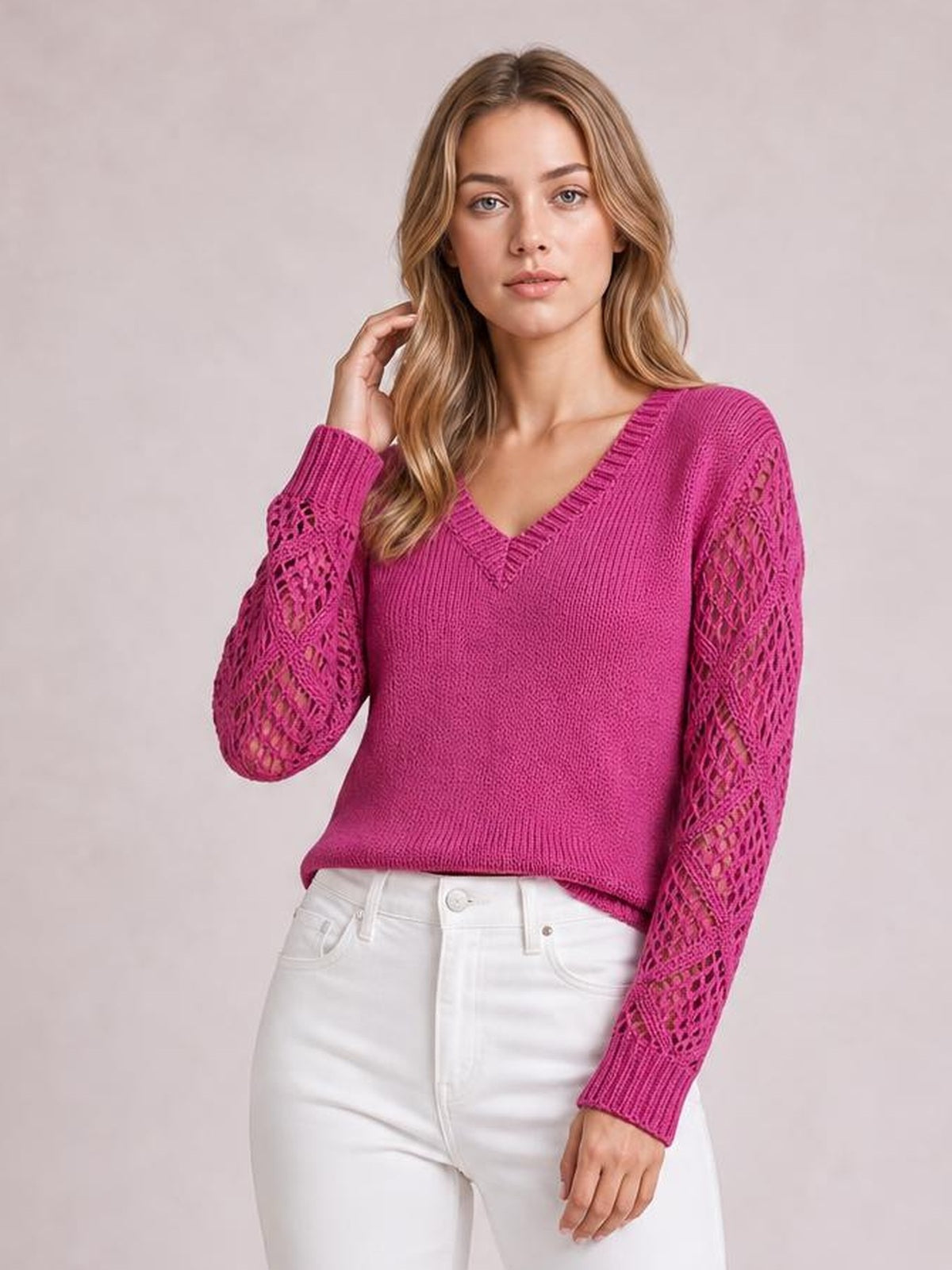 Model wearing a bright pink V-neck knit sweater with openwork hollow-out sleeves, ribbed trim, and a relaxed silhouette, posing with one hand near her hair.