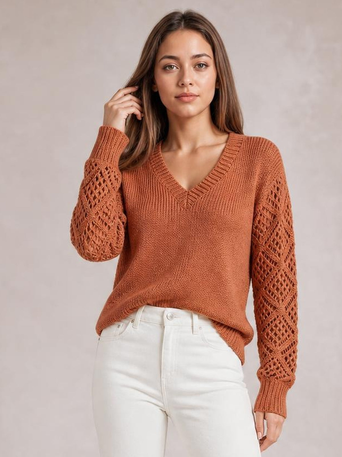 Model poses in a rust V-neck knit sweater with openwork hollow-out sleeves, ribbed trim, and a relaxed silhouette over white jeans.