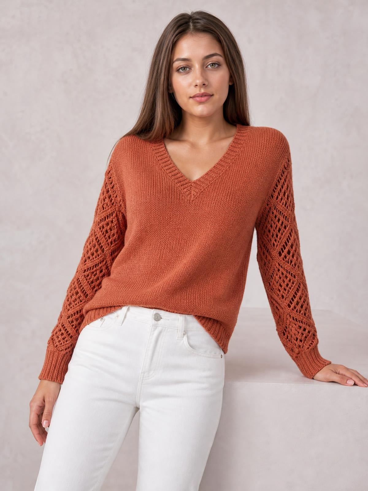 Model in a rust V-neck knit sweater with openwork hollow-out sleeves, ribbed trim, a relaxed silhouette, and one hand resting on a ledge.