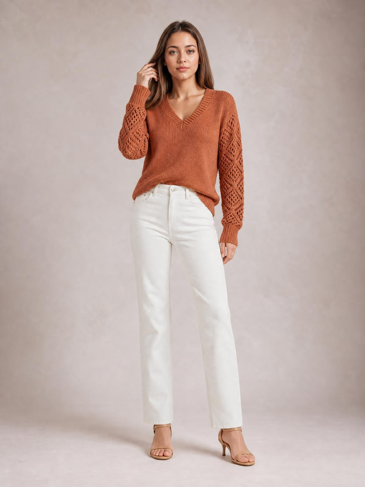 Model wearing a rust V-neck knit sweater with openwork hollow-out sleeves, ribbed trim, and a relaxed straight silhouette, posing with one hand in her hair.