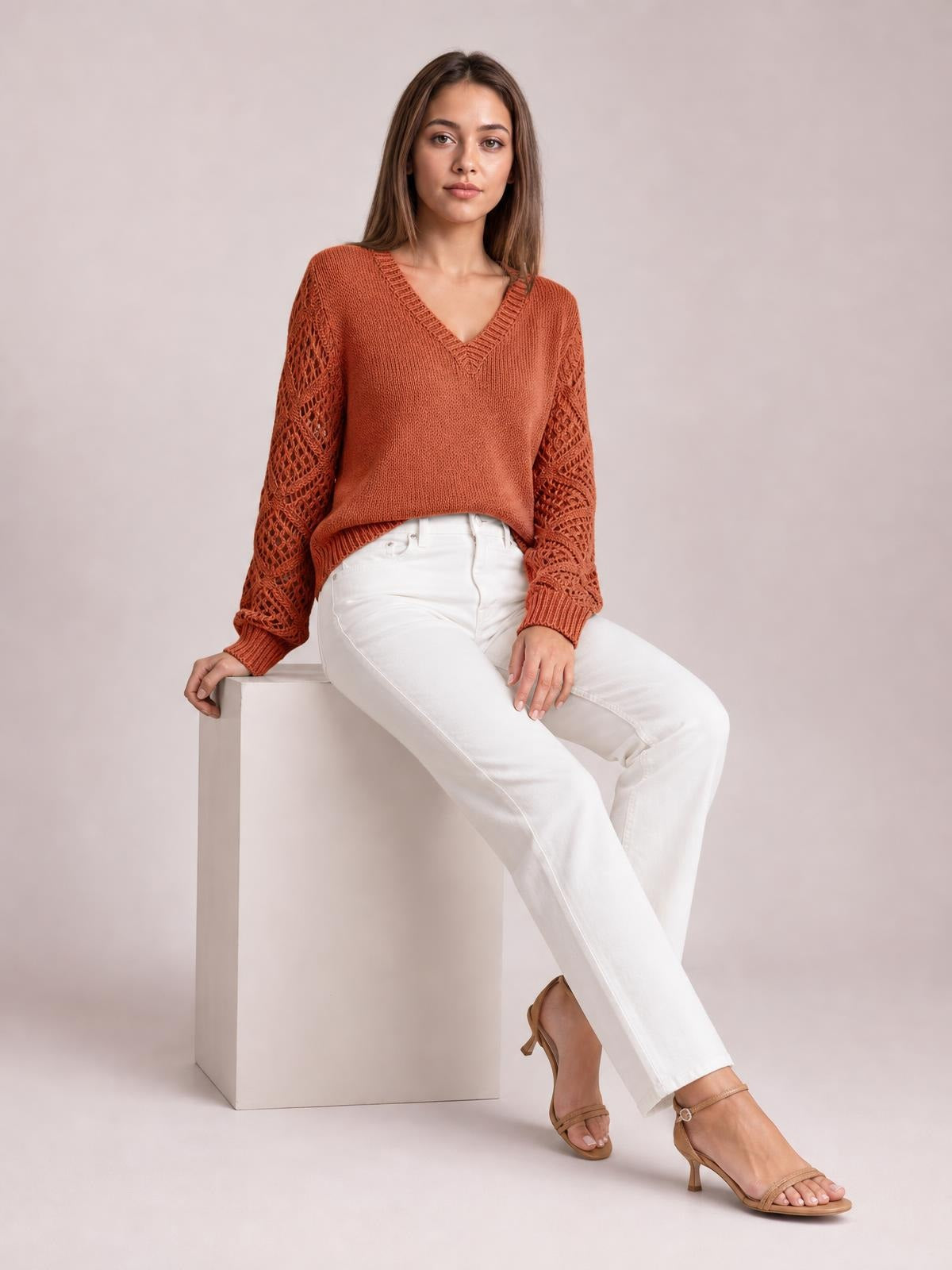 A model sits wearing a rust-orange V-neck knit sweater with openwork hollow-out sleeves, ribbed cuffs and hem, and a relaxed silhouette.