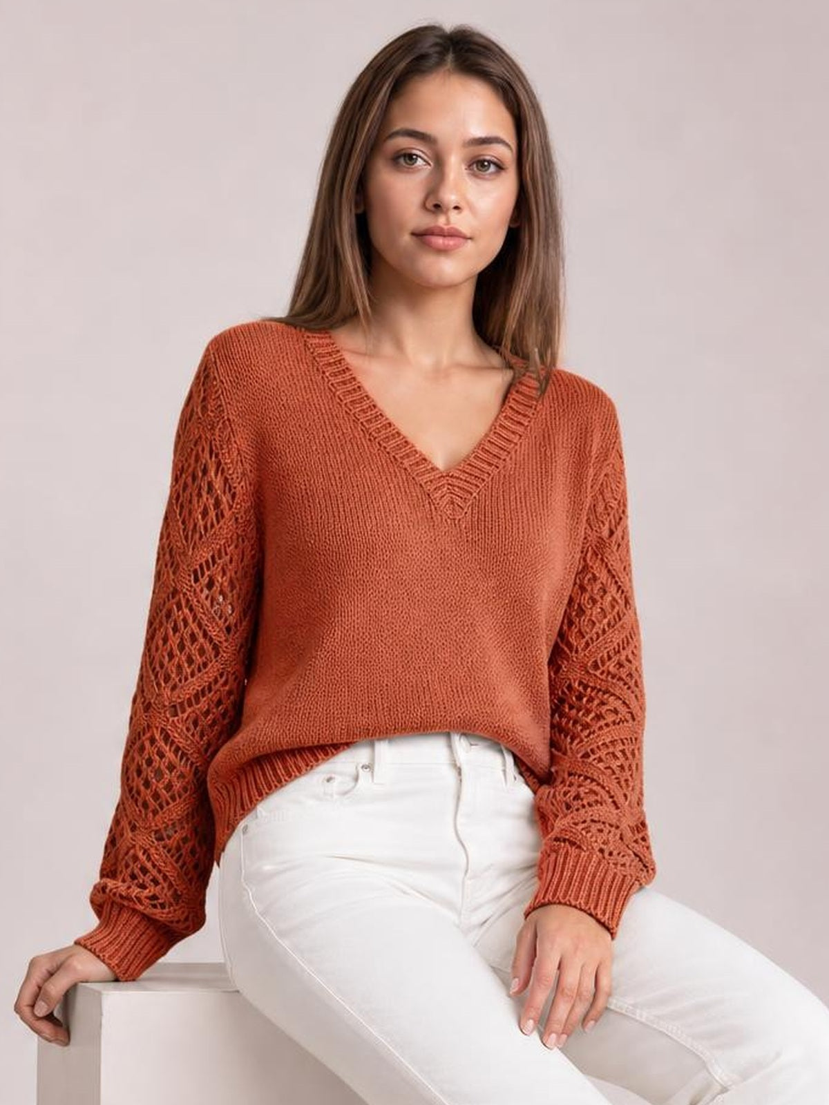 Model seated in a rust V-neck knit sweater with openwork hollow-out sleeves, ribbed trim, long sleeves, and a relaxed silhouette.