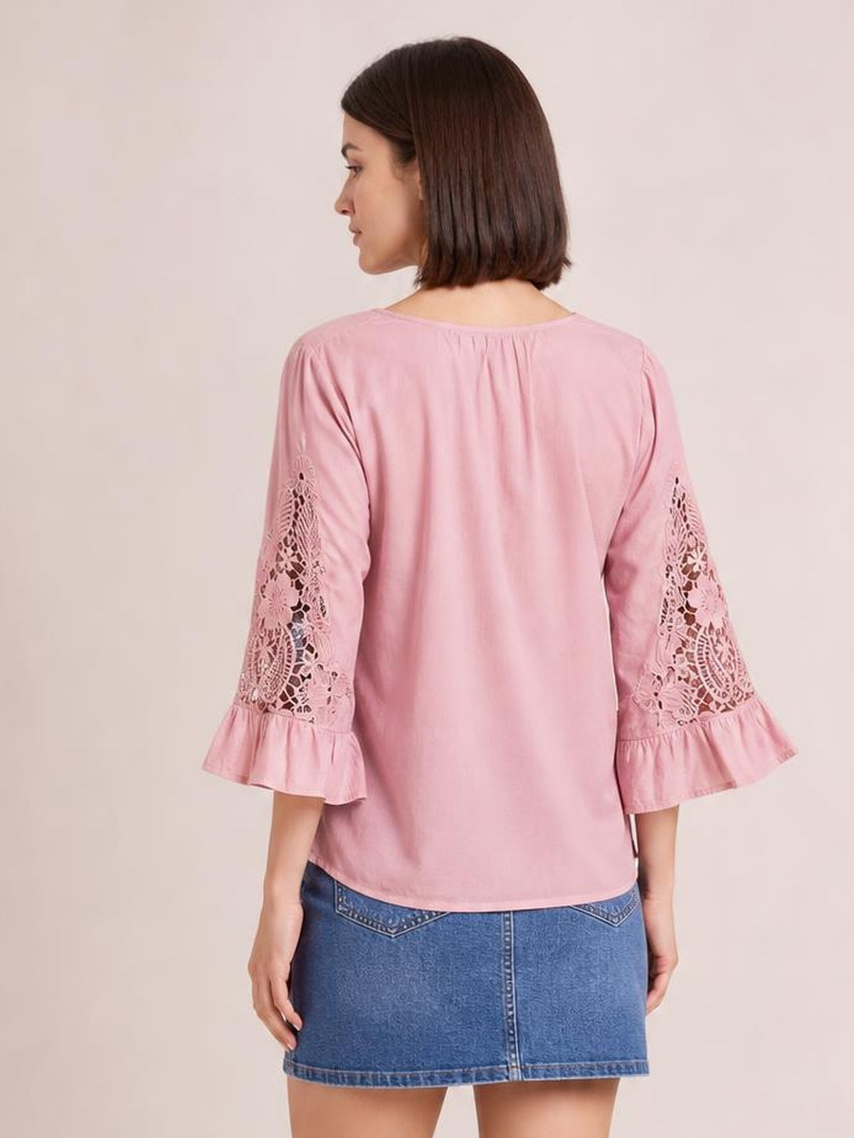 A model stands facing away in a pink blouse with a relaxed silhouette, three-quarter lace-inset sleeves, and flounce cuffs.