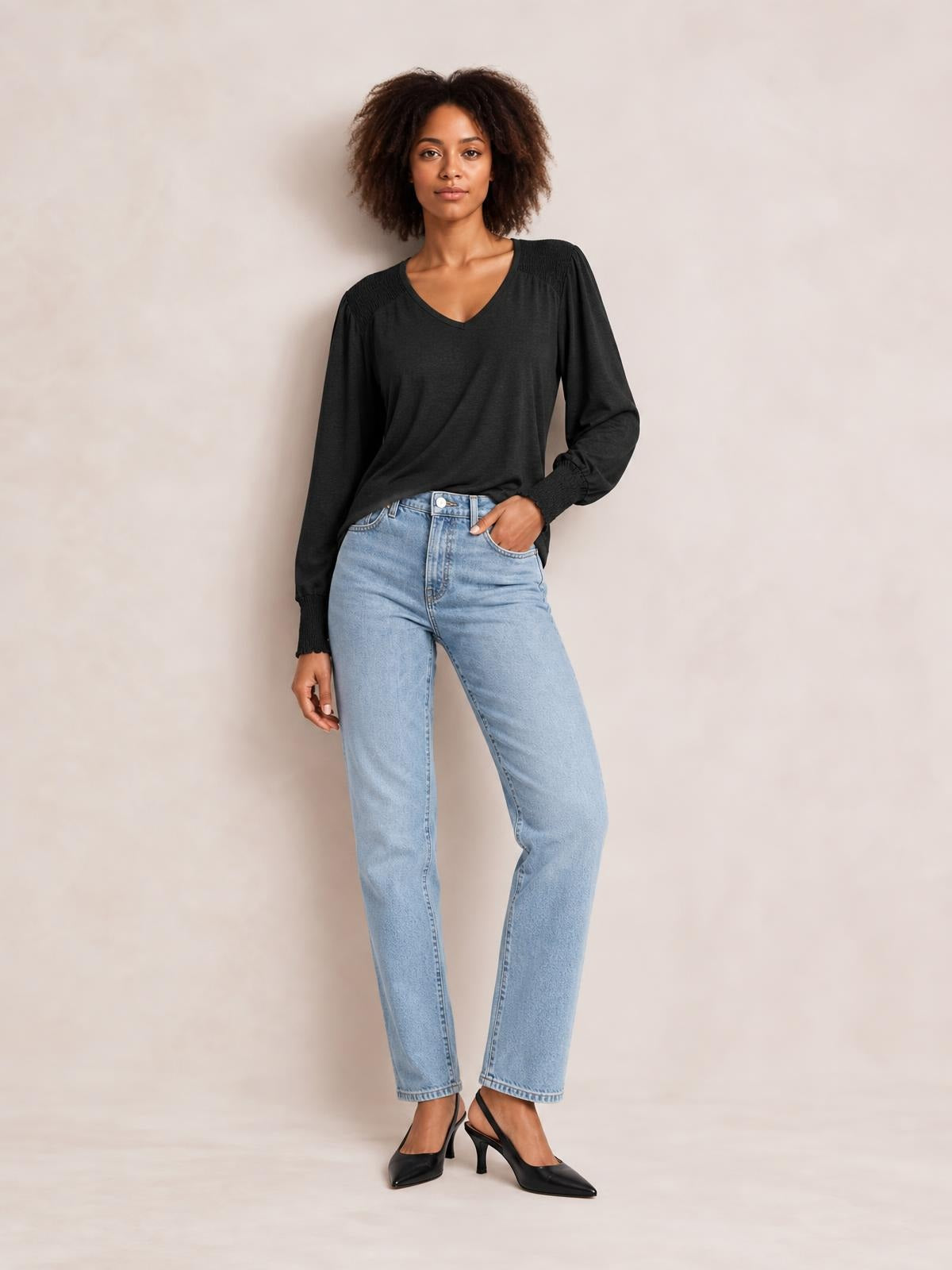 Model wears a black V-neck lantern sleeve T-shirt with a relaxed silhouette, gathered shoulder detail, and smocked cuffs, posing with one hand in her jeans pocket.