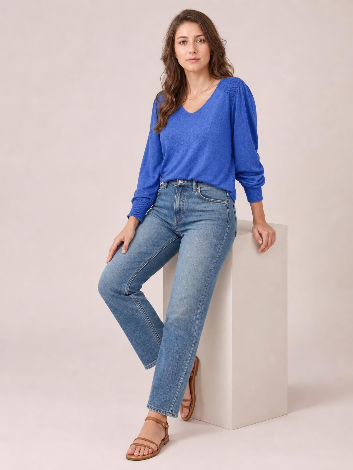 Model seated with one leg crossed, wearing a blue V-neck T-shirt with voluminous lantern sleeves, gathered cuffs, and a relaxed straight silhouette.