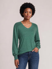 Model wears a forest-green V-neck lantern sleeve T-shirt with a relaxed silhouette, gathered shoulders, and ribbed cuffs, standing with one hand in her jeans pocket.