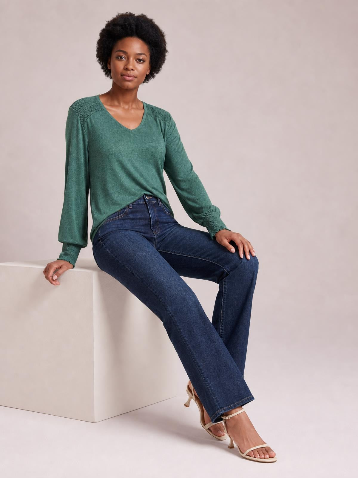 A seated model wears a forest-green V-neck T-shirt with long lantern sleeves, gathered cuffs, and a relaxed silhouette styled with jeans.