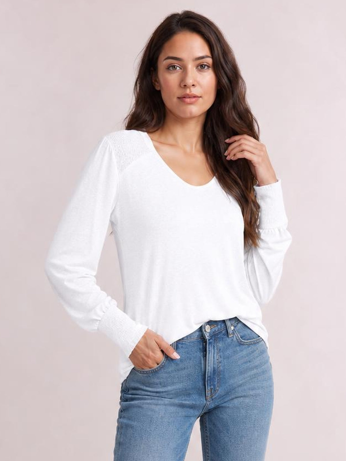 Model poses in a white V-neck T-shirt with long lantern sleeves, gathered cuffs, and a relaxed silhouette, touching her hair with one hand.