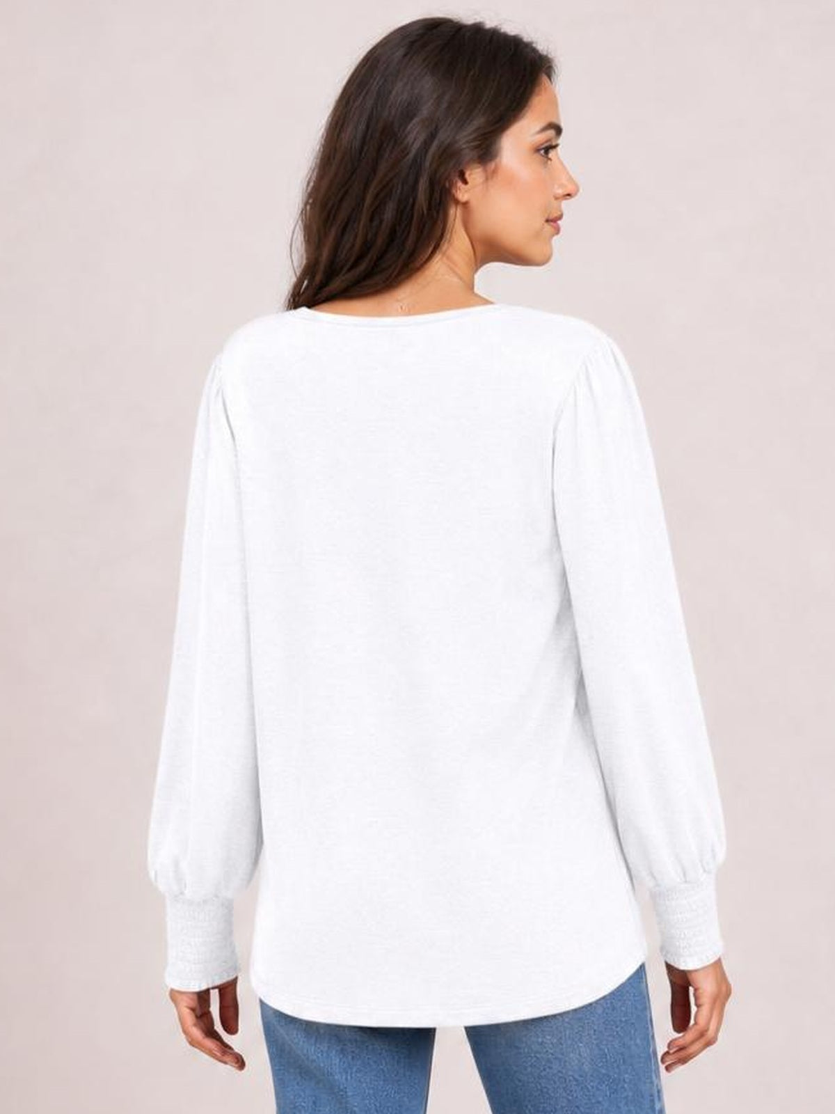A model turns to show the back of a white V-neck T-shirt with long lantern sleeves, gathered cuffs, a relaxed silhouette, and a gently curved hem.