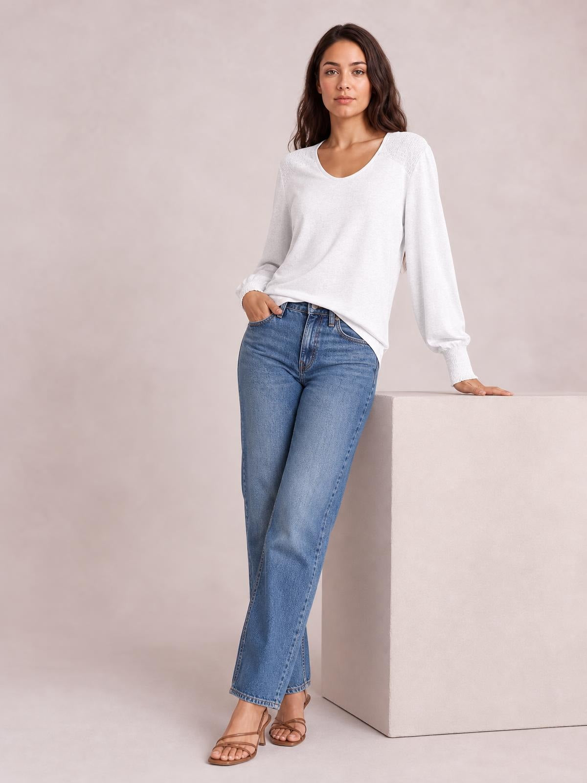 A model wears a white long-sleeve T-shirt with lantern sleeves, gathered cuffs, a relaxed silhouette, and a gently curved neckline while leaning on a block.