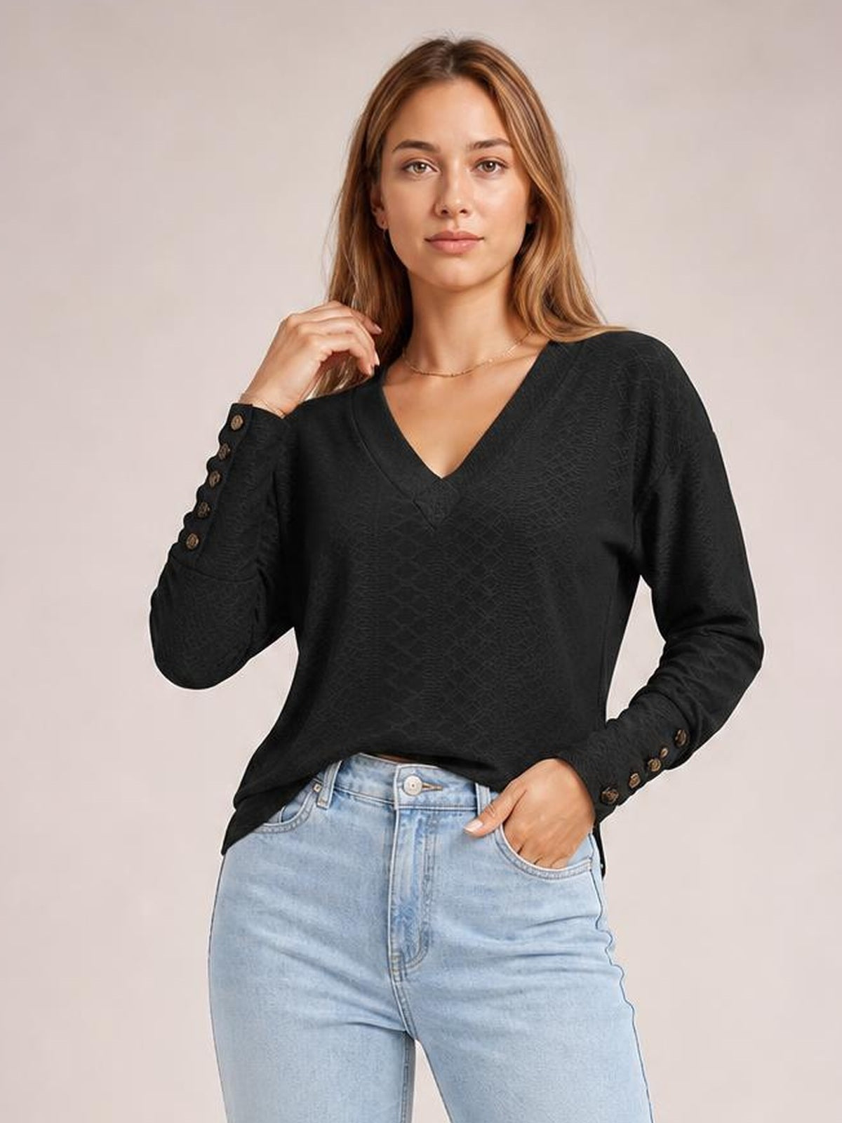 Model wearing a black V-neck long sleeve blouse with a regular silhouette, subtle texture, and button-accented cuffs, posing with one hand in her jeans pocket.