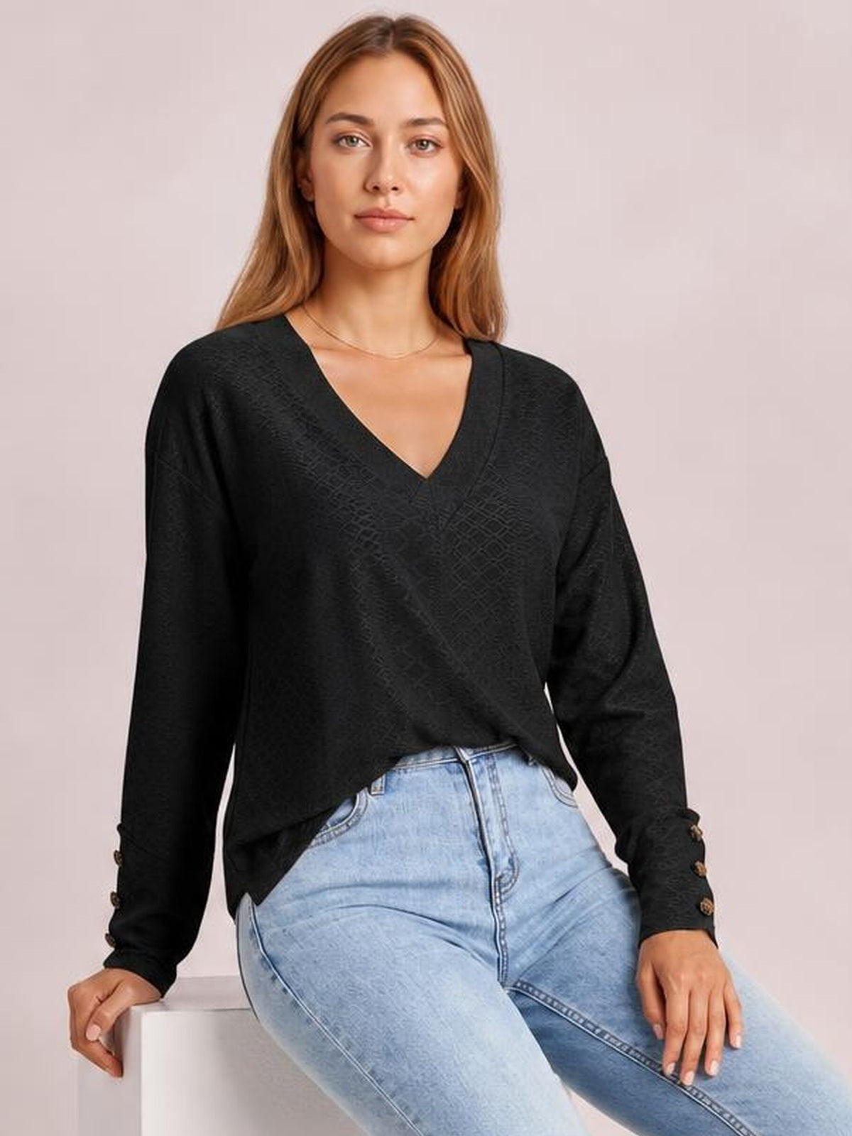 Model wearing a solid black V-neck long sleeve blouse with a relaxed regular silhouette and button-accented cuffs, seated with one hand resting on a block.