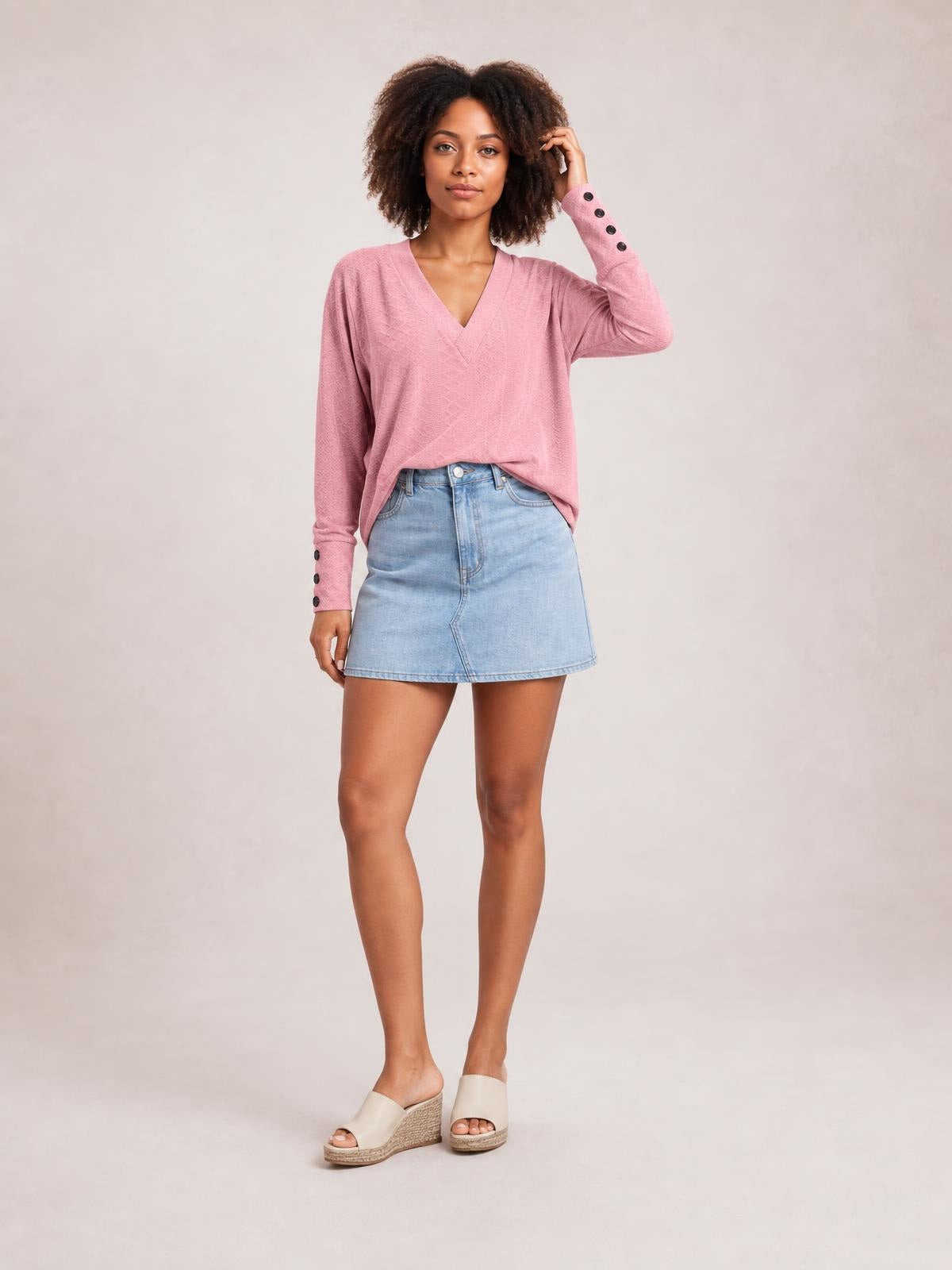 Model wearing a solid pink V-neck long sleeve blouse with a regular relaxed silhouette and buttoned cuffs, posing with one hand touching her hair.