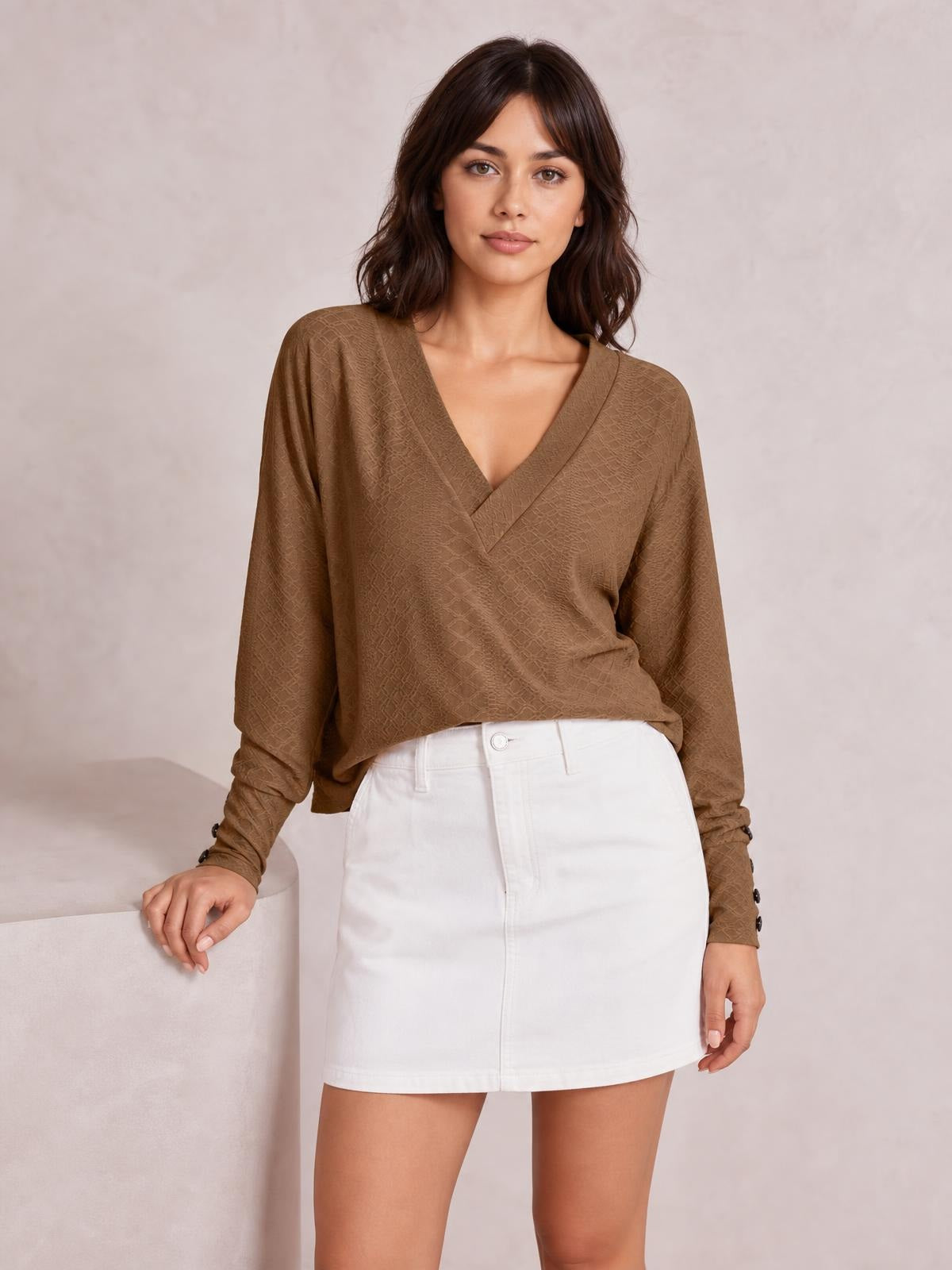 A model poses in a brown V-neck long sleeve blouse with a relaxed regular silhouette, subtle woven texture, and buttoned cuffs, resting one hand on a ledge.