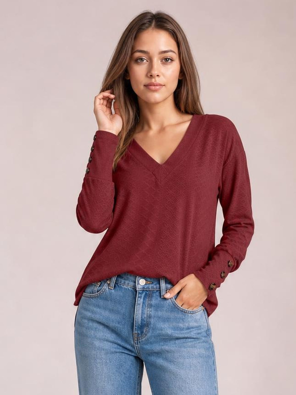 Model wears a burgundy V-neck long sleeve blouse with a relaxed regular silhouette, subtle tonal texture, and buttoned cuffs, posed with one hand in her jeans pocket.