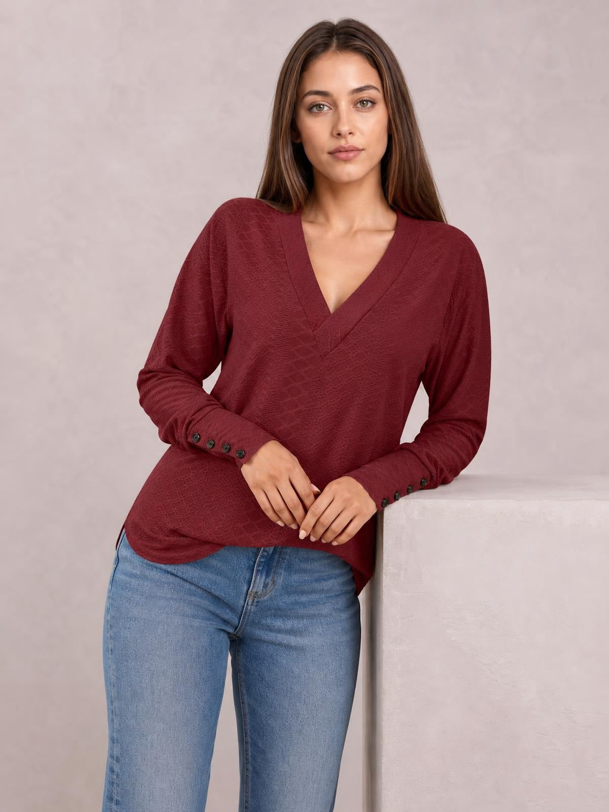Model wears a burgundy V-neck long sleeve blouse with a relaxed regular silhouette and buttoned cuffs, posing with hands resting in front.