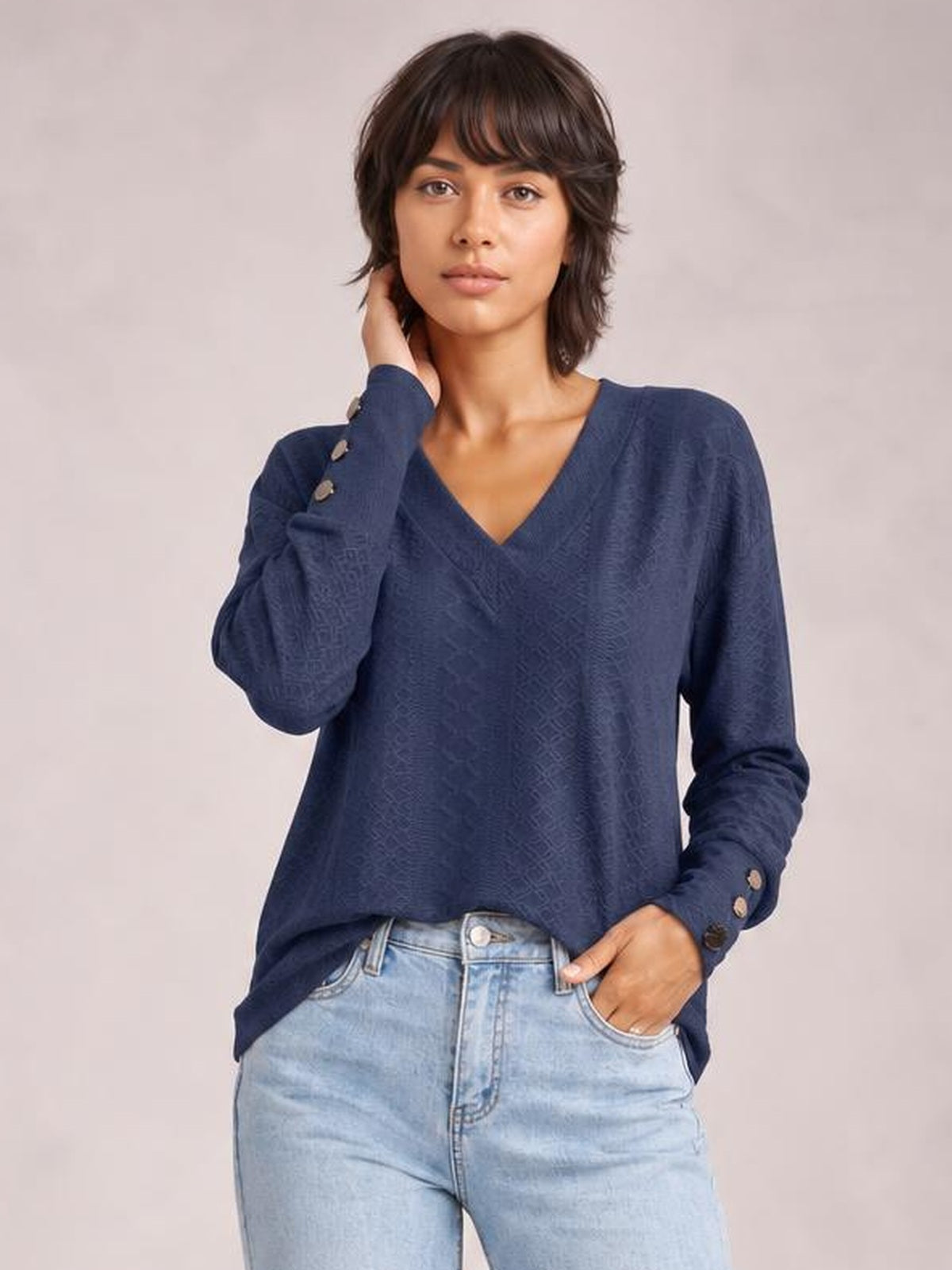 Model wearing a solid navy V-neck long sleeve blouse with a relaxed regular-length silhouette and button-detail cuffs, posing with one hand in her jeans pocket.