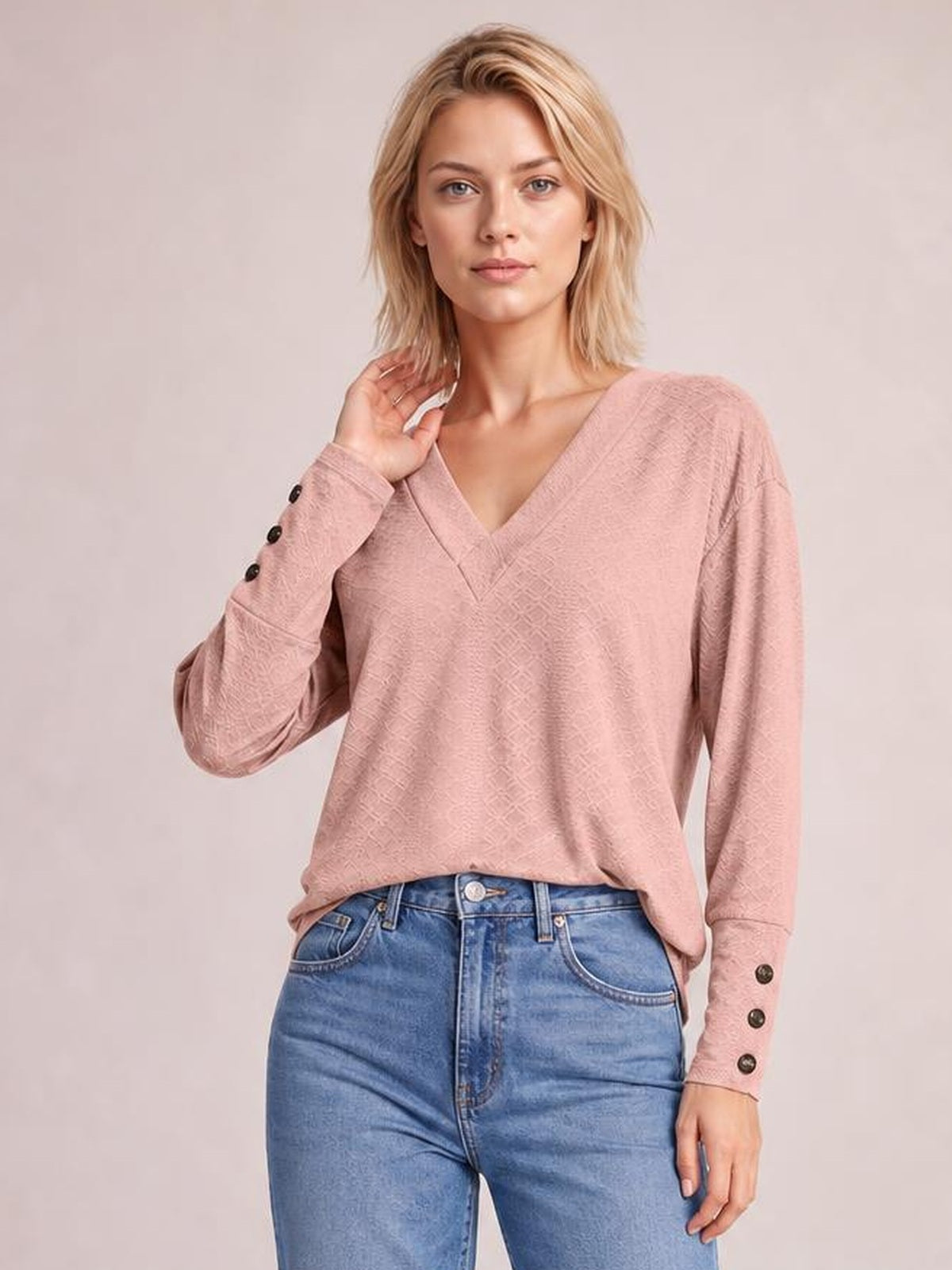 Model wears a solid blush pink V-neck long sleeve blouse with a relaxed regular silhouette, buttoned cuffs, and a slightly tucked hem with one hand touching her hair.