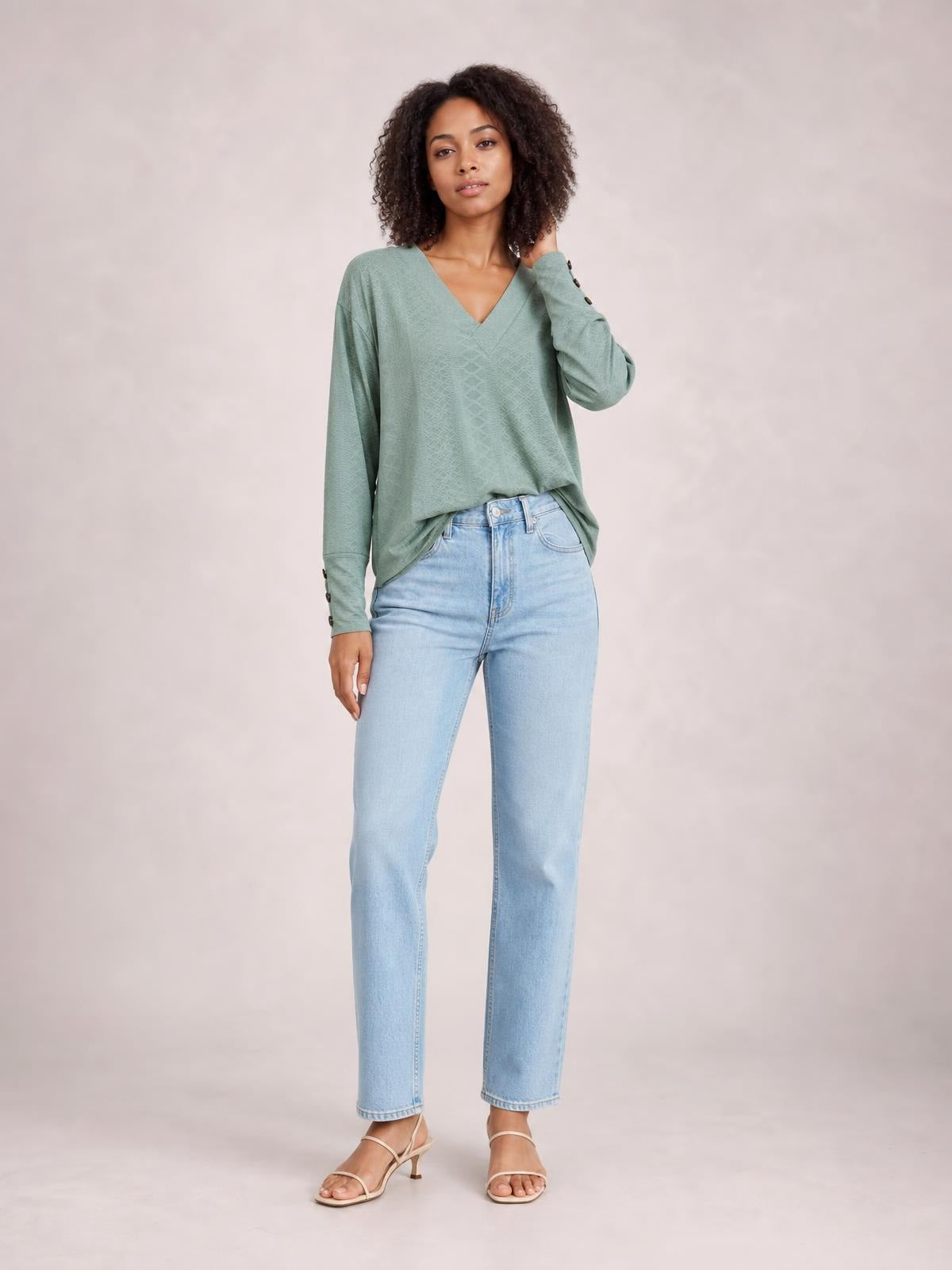 Model wearing a sage green V-neck long sleeve blouse with a relaxed regular-length silhouette and button cuff details, posing with one hand near her hair.