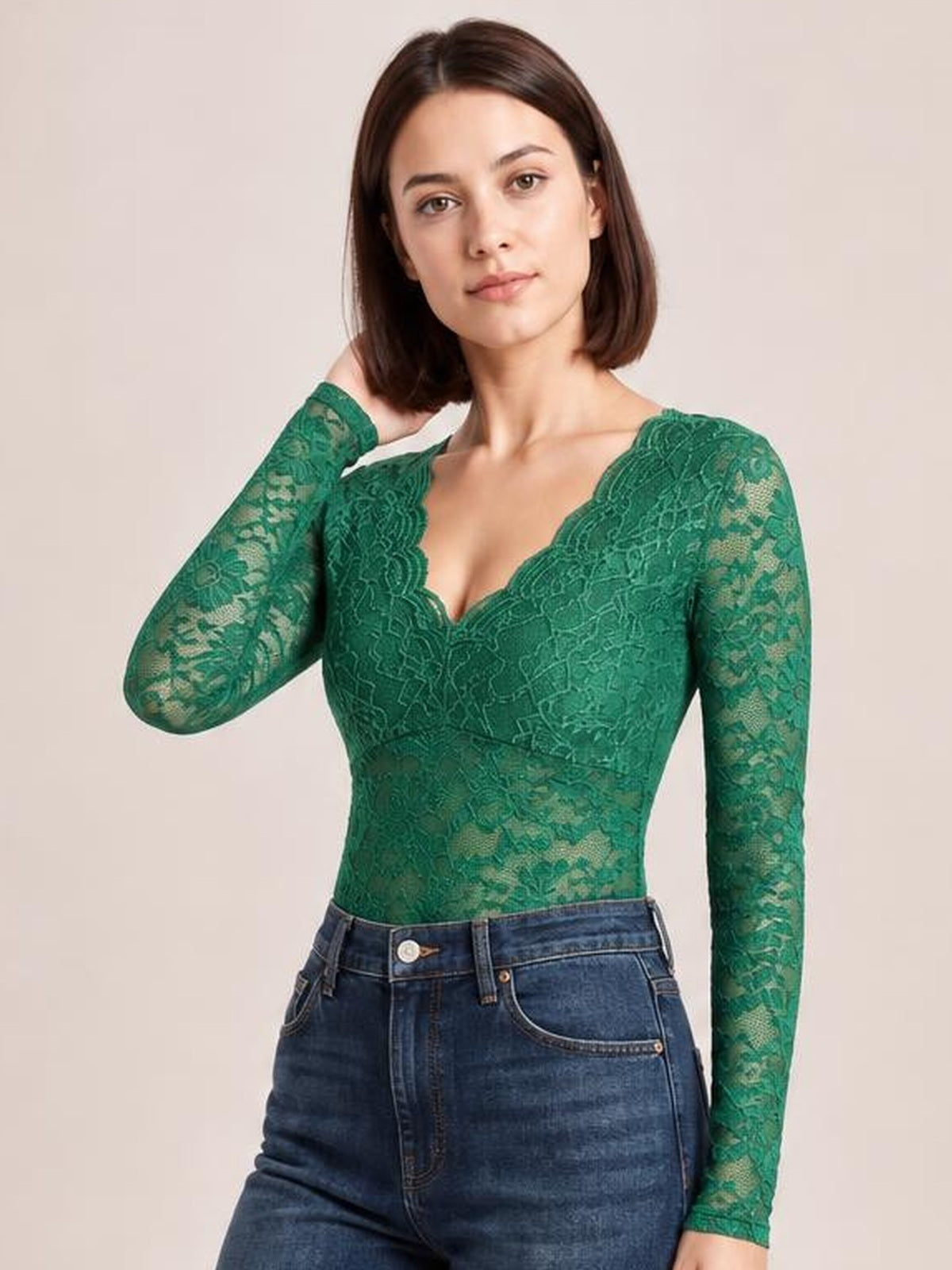 Model poses with one hand behind her neck, wearing a fitted green V-neck long sleeve lace top with scalloped trim and sheer lace sleeves.