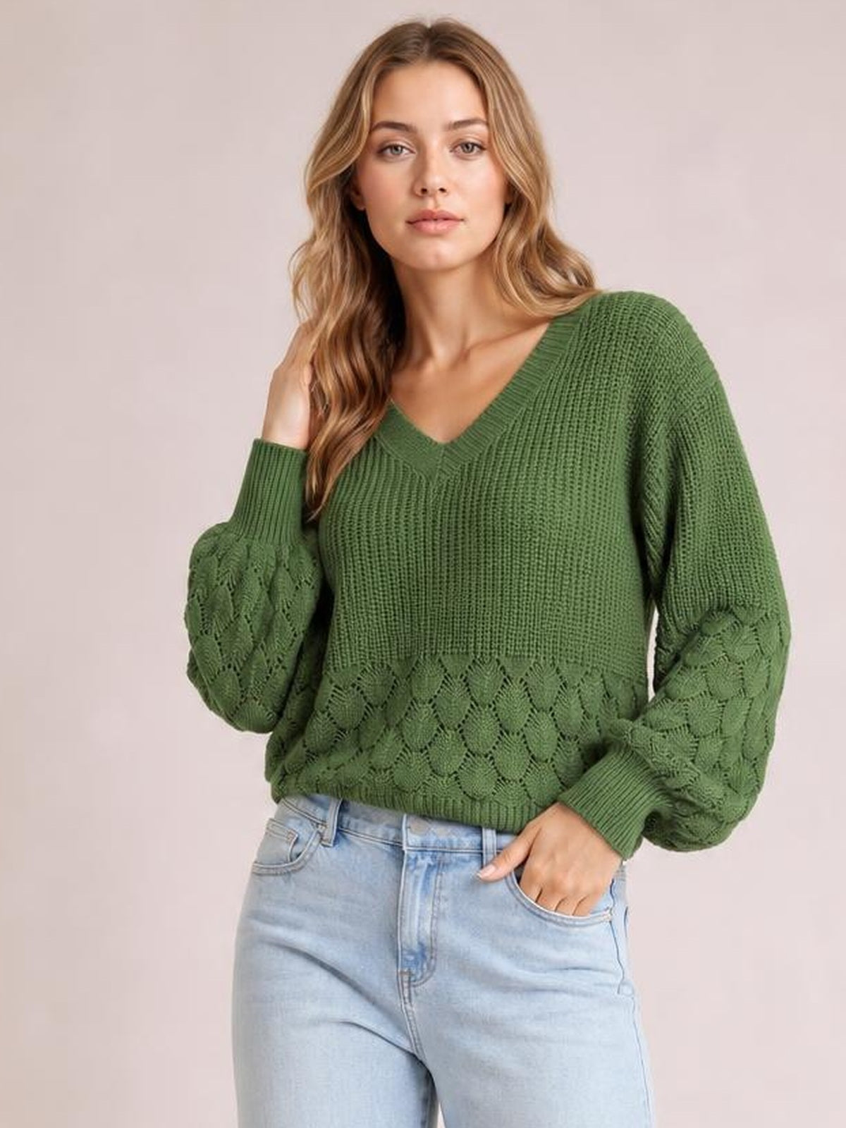 Model wearing a relaxed green V-neck long sleeve sweater with textured knit panels, voluminous sleeves, ribbed cuffs and hem, paired with light-wash jeans.