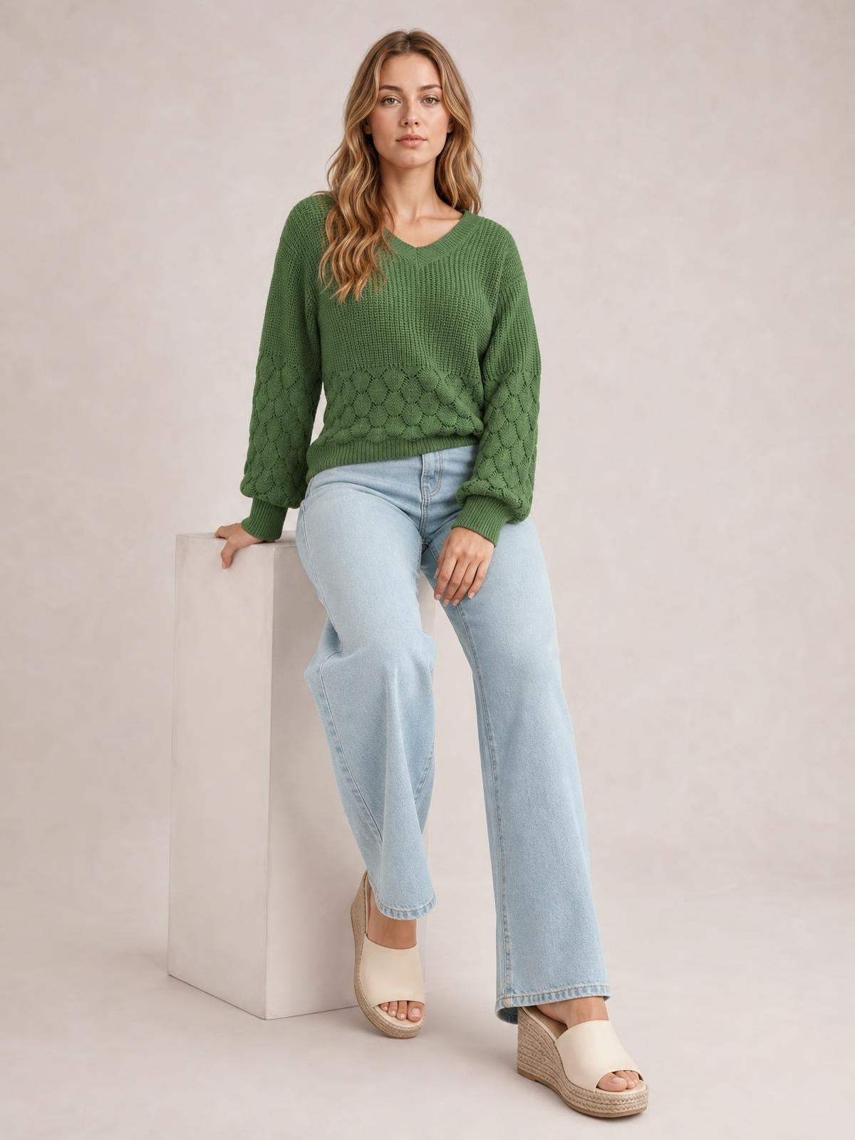 Model seated in a relaxed pose wearing a green V-neck long sleeve sweater with a loose silhouette, ribbed cuffs and hem, and textured knit panels.