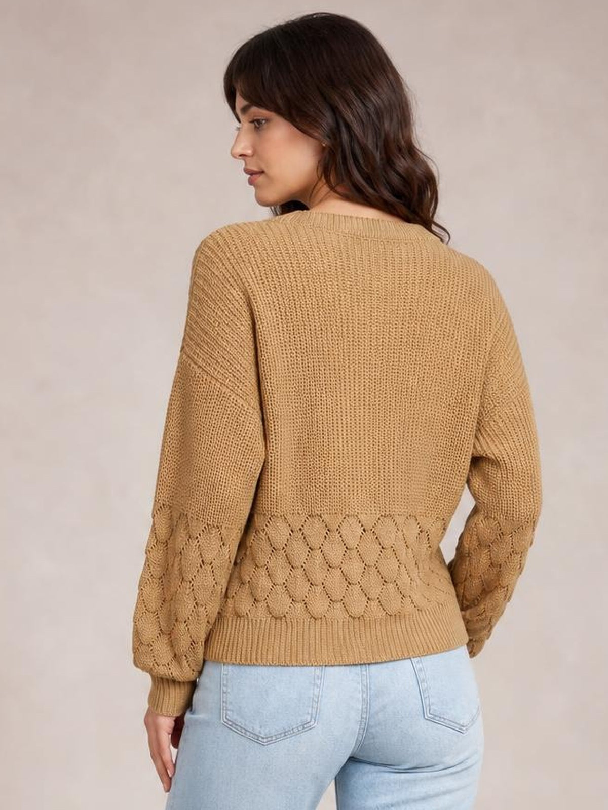 Model looks over her shoulder in a camel V-neck long sleeve sweater with a relaxed fit, ribbed trim, and diamond-knit detailing across the lower back and sleeves.