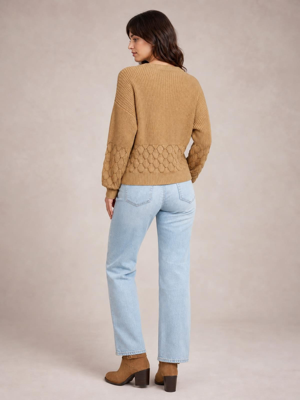 A model turned away wears a relaxed tan long-sleeve sweater with textured knit construction, openwork detailing at the lower sleeves and hem, and ribbed trim.