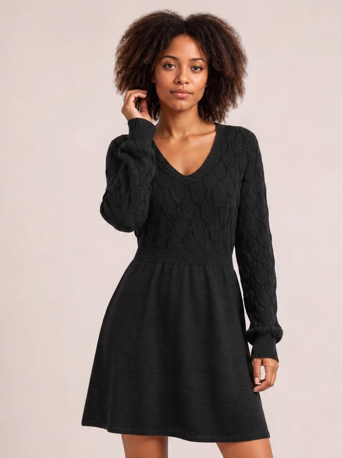 A model poses in a black V-neck long sleeve sweater dress with openwork knit detailing, a fitted waist, ribbed cuffs, and a softly flared above-knee silhouette.