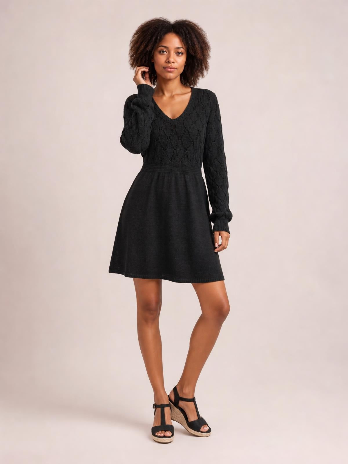 A model poses in a black V-neck long sleeve sweater dress with openwork knit sleeves and bodice, a defined waist, and a softly flared knee-length skirt.