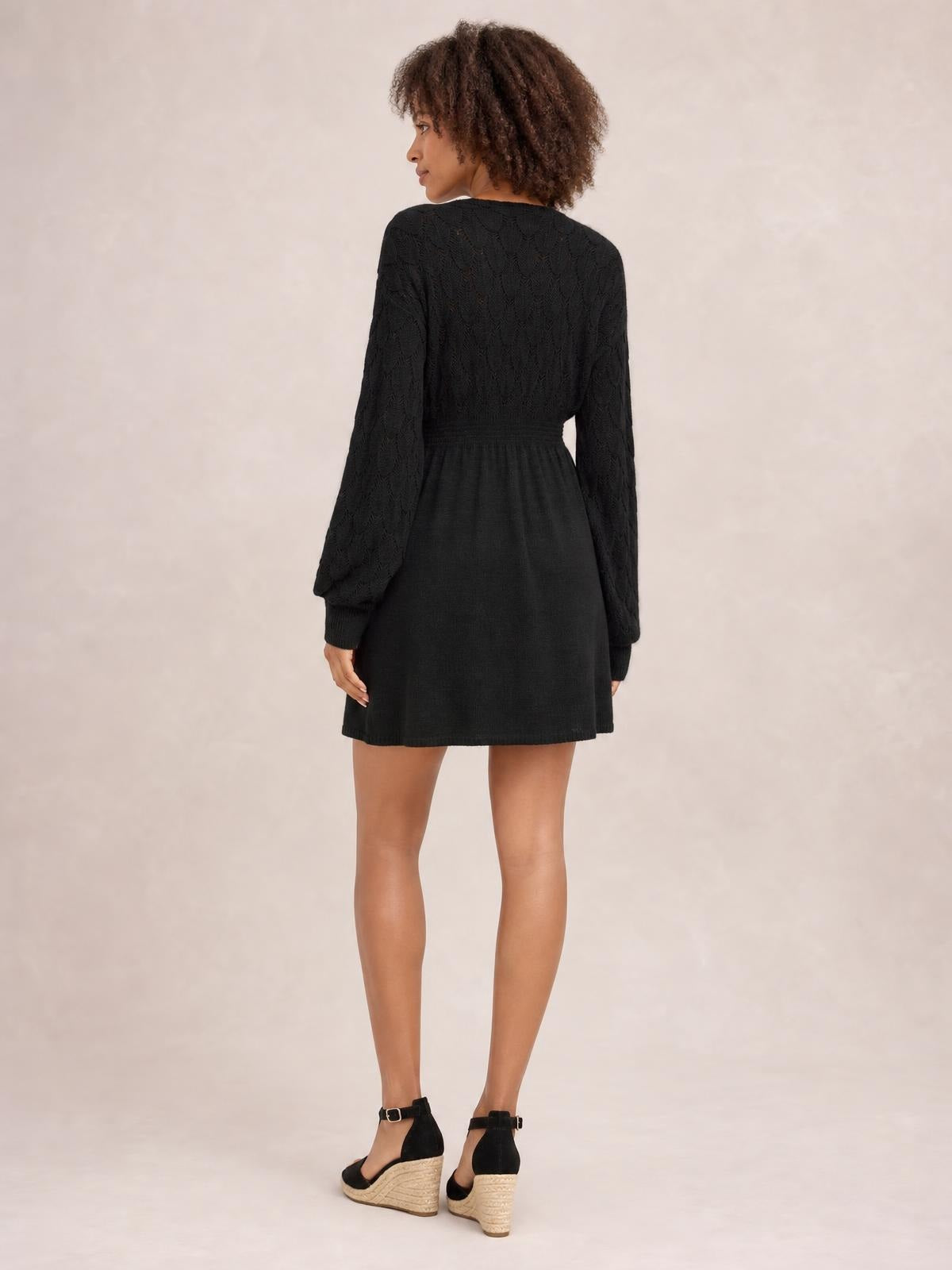 A model poses in a black openwork long-sleeve sweater dress with a defined waist and softly flared short skirt, shown from the back.