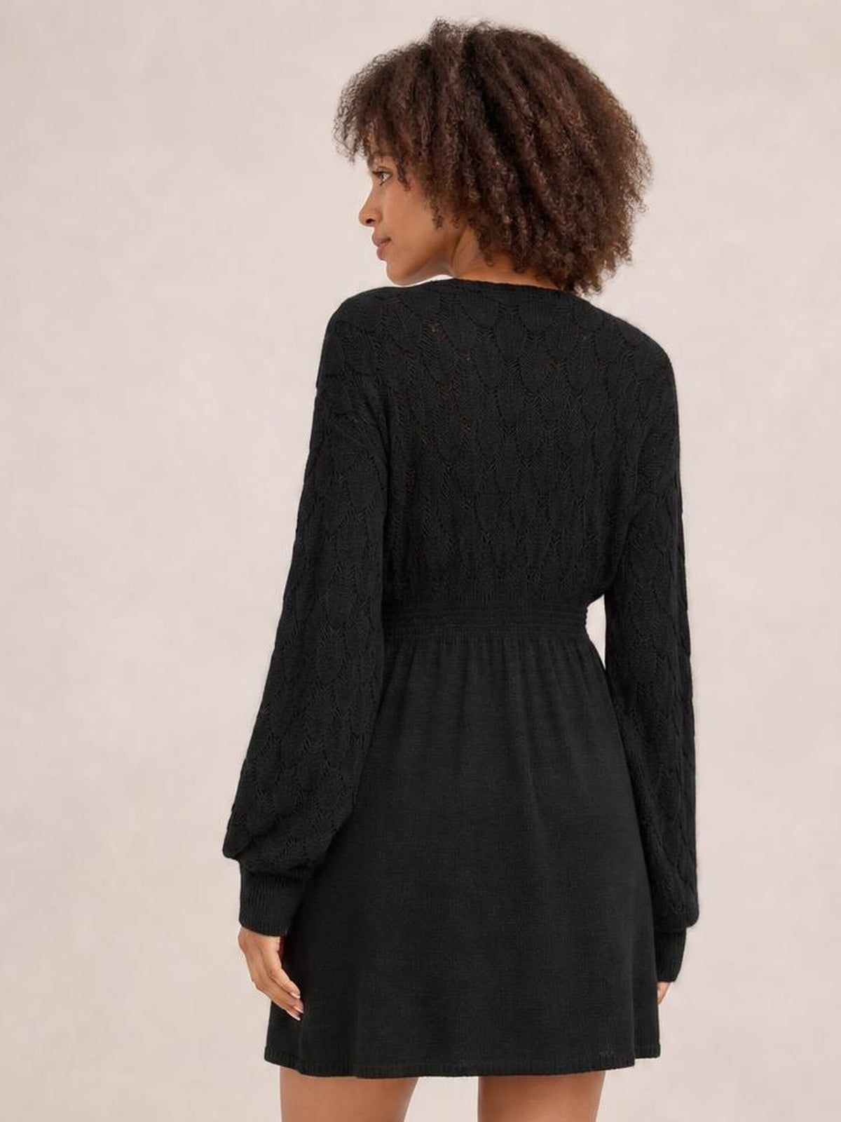 Model shown from the back in a black long sleeve sweater dress with openwork knit, a defined waist, ribbed cuffs, and a gently flared above-knee skirt.