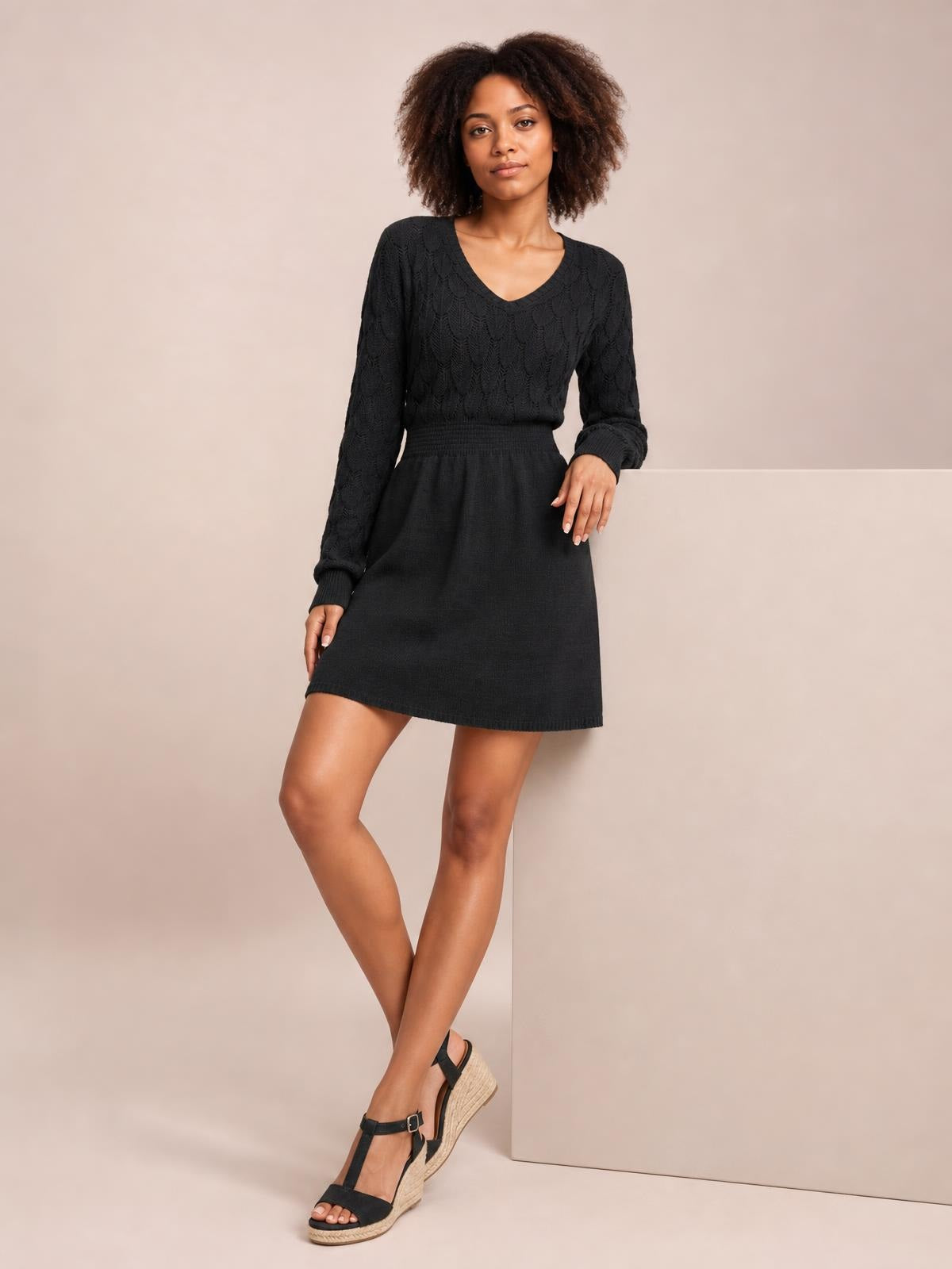 A model wears a black V-neck long-sleeve sweater dress with openwork knit texture, fitted bodice, ribbed waist, and flared mini silhouette while posing with crossed legs.