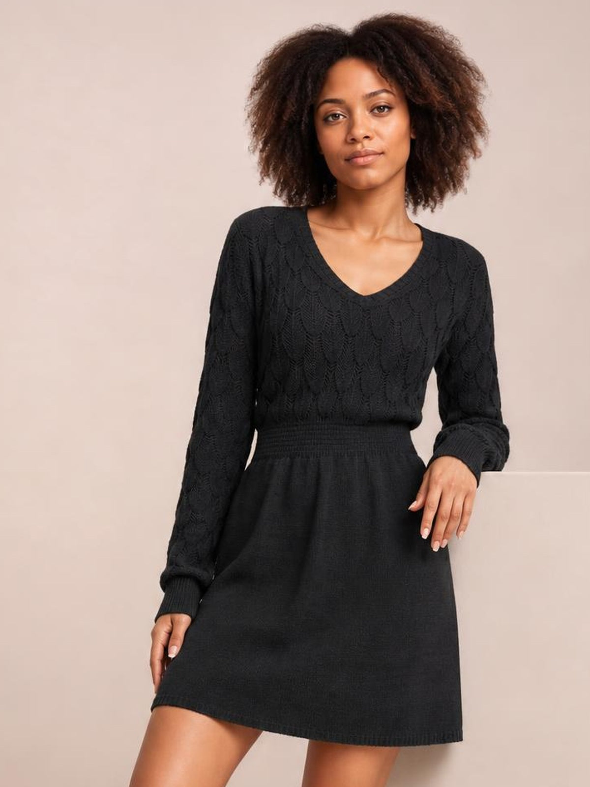 Model poses in a black V-neck long sleeve sweater dress with openwork knit texture, ribbed cuffs and waistband, a fitted bodice, and gently flared short hem.