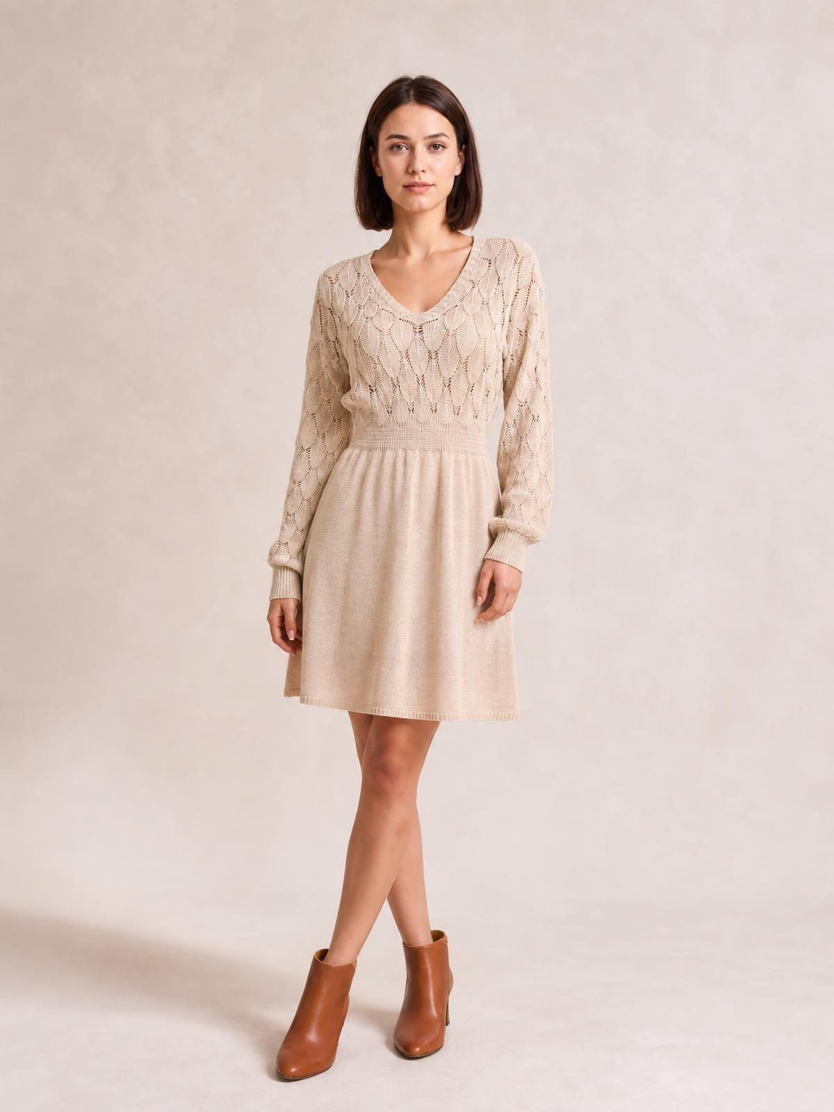 Model stands with crossed legs in a cream V-neck long sleeve sweater dress featuring openwork knit, ribbed cuffs and waist, and a softly flared above-knee silhouette.