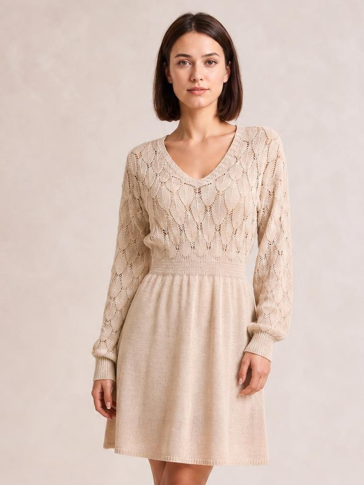 Model stands facing forward in a beige V-neck long sleeve sweater dress with openwork knit detailing, ribbed cuffs and waistband, and a gently flared silhouette.