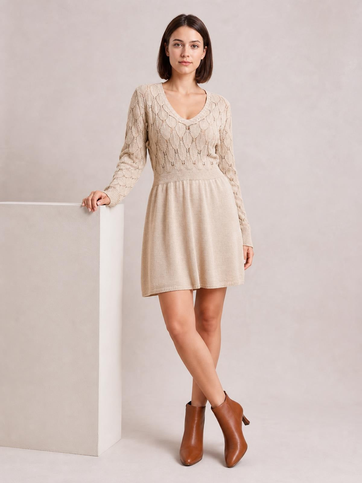 A model poses with one hand on a pedestal in a beige openwork V-neck long sleeve sweater dress with a fitted waist and flared mini silhouette.