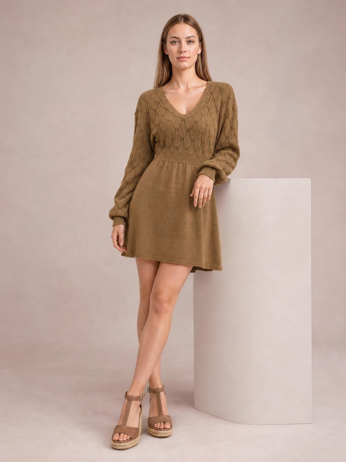 A model stands with crossed legs in a taupe V-neck long sleeve sweater dress featuring openwork knit detailing, a defined waist, and a flared mini silhouette.
